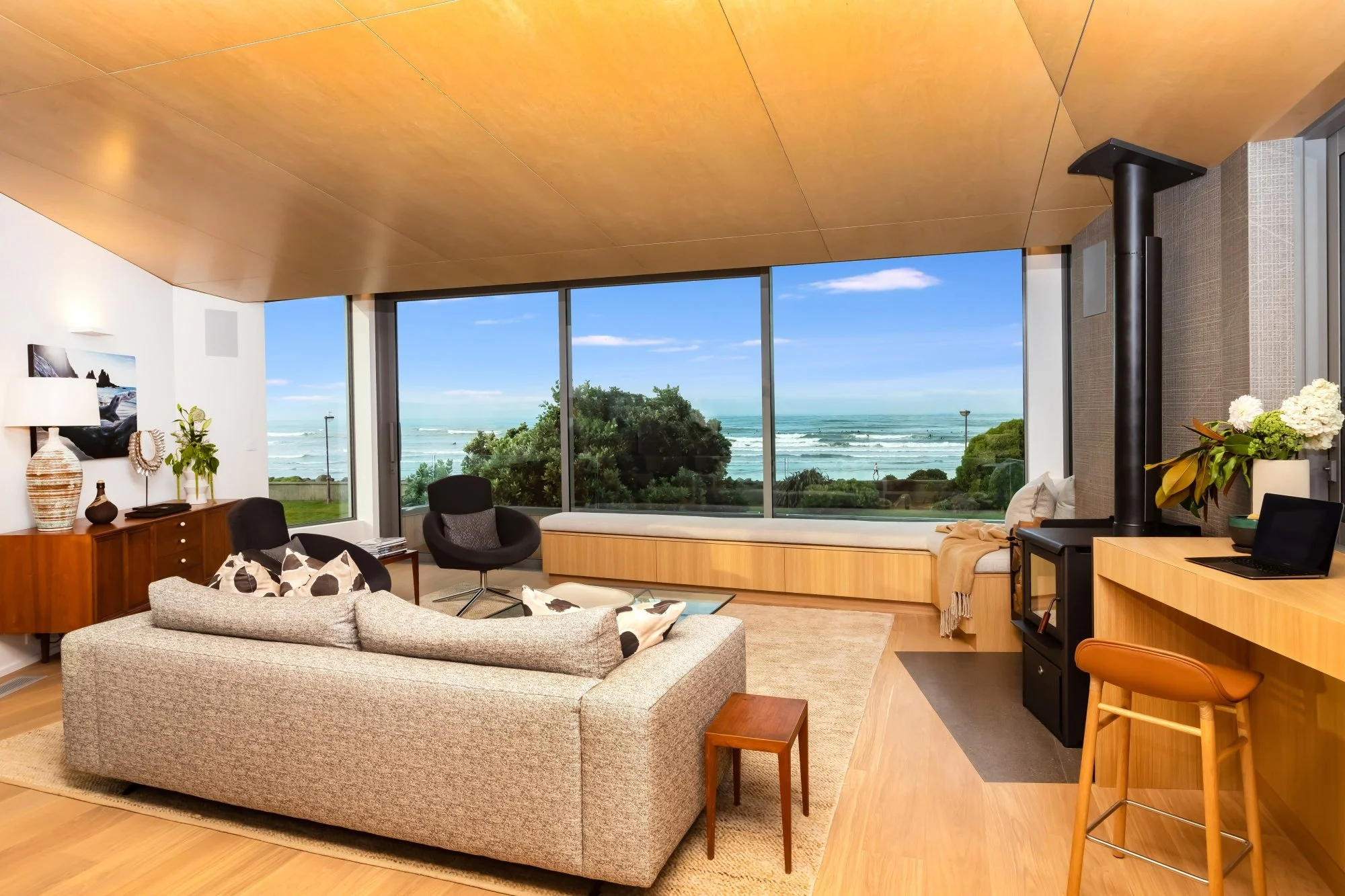 Living room with large window view of ocean and sky, beige and wooden furniture, a sofa, armchairs, a side table, a cabinet with decorative items, a television, a wood stove, and a plant.
