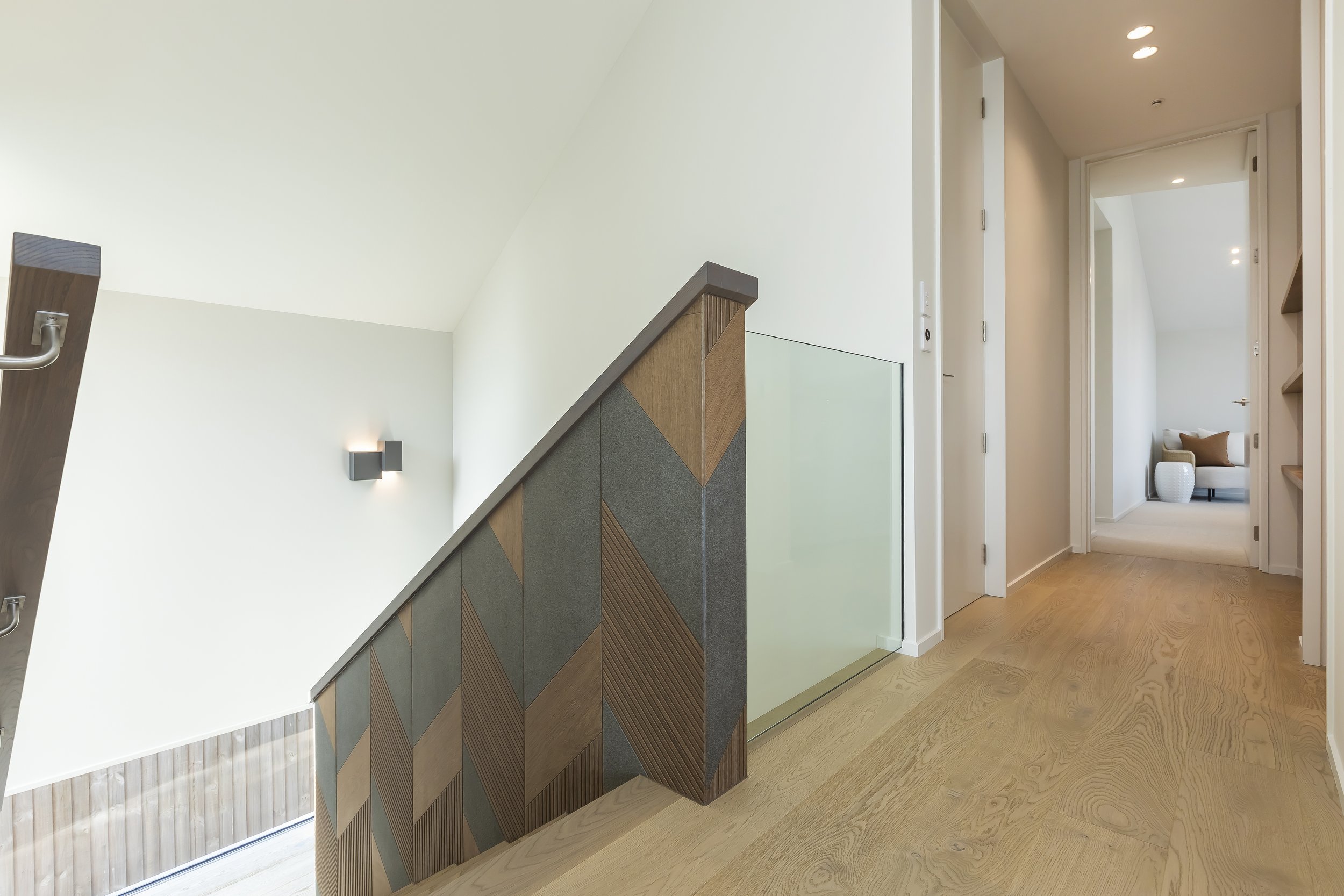 Interior of a modern home showing a staircase with a wooden and geometric design, beige hardwood floors, white walls, and hallways leading to other rooms with visible furniture. Stair Design by Bridget Coles Interior Design