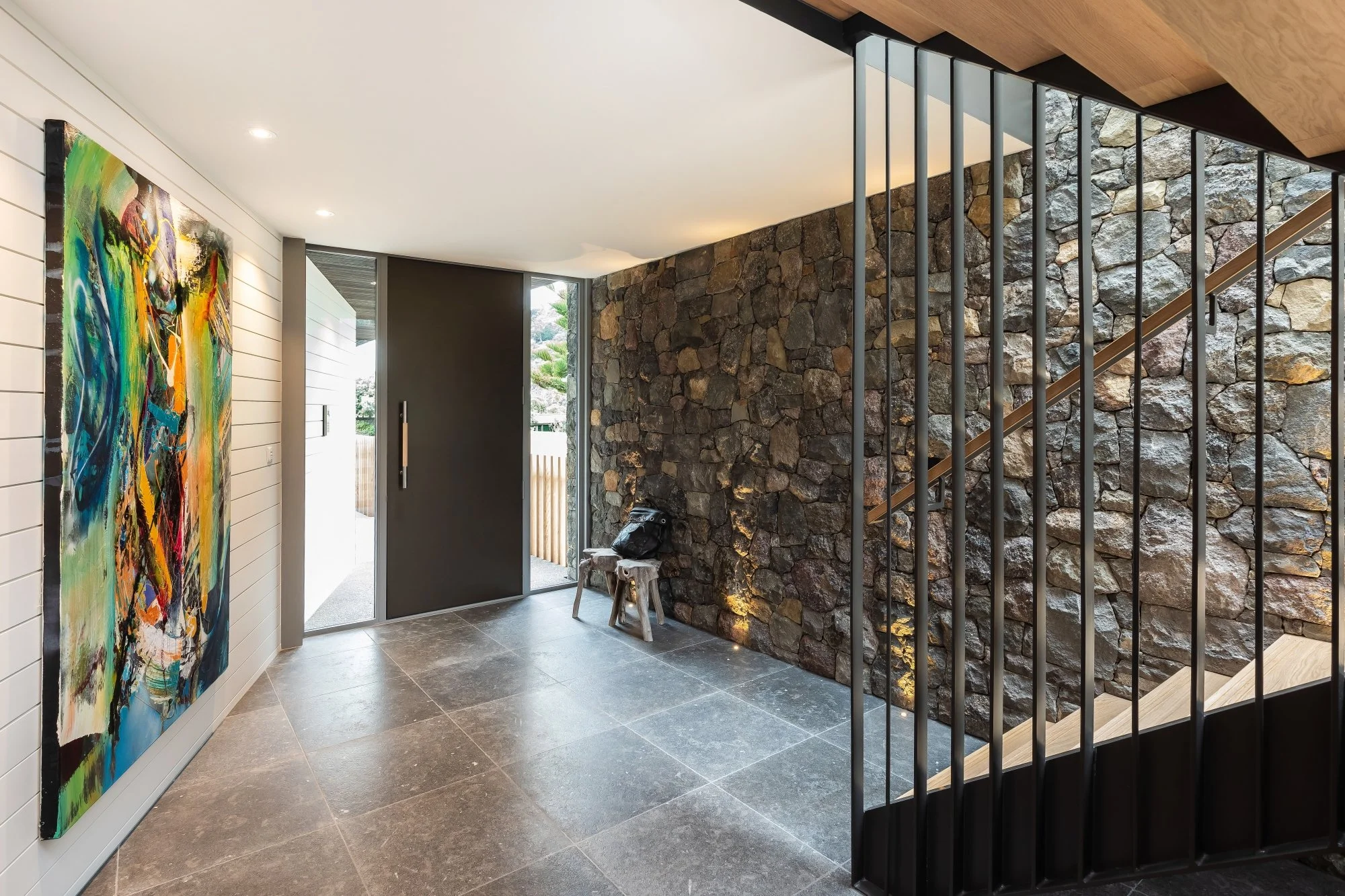 Modern interior entryway featuring a Dulux powdercoated door, a large colorful abstract painting on white shiplap wall, a stone accent wall, Chamboille gris flooring, and a staircase with black metal railing.