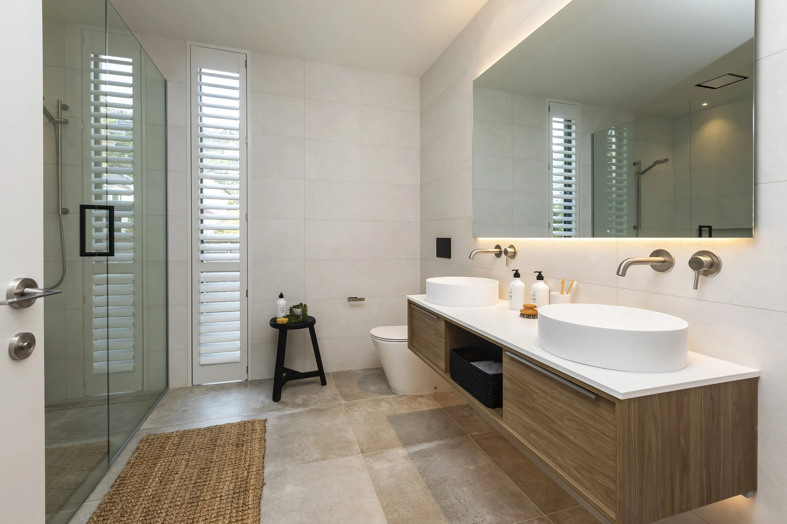 A modern bathroom with a walk-in shower with glass doors on the left, a large mirror above a double sink vanity on the right, white walls, and beige floor tiles. Tall windows with Sante Fe shutters are visible, and there is a small black stool with t