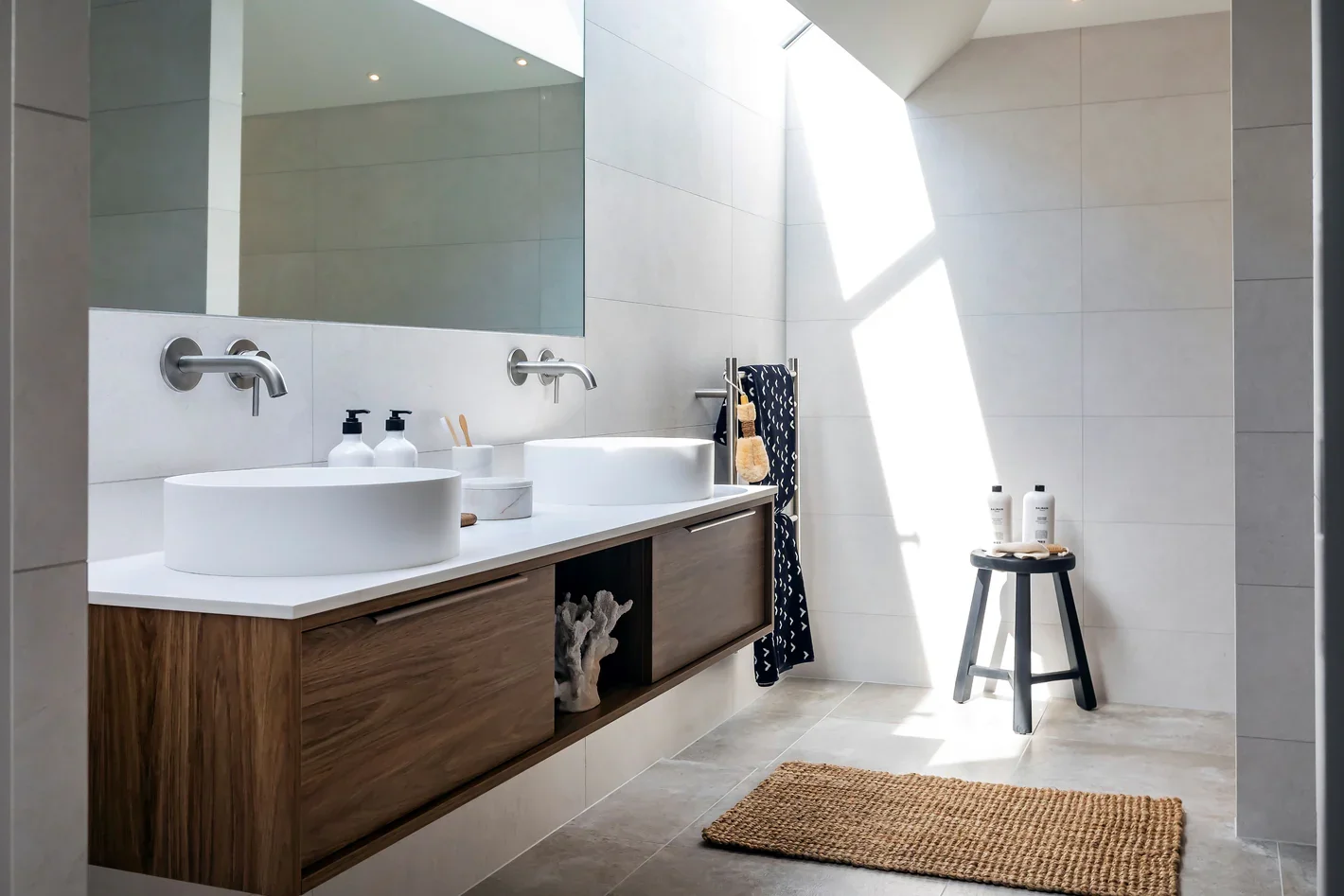 Modern bathroom with two vessel sinks and a large mirror, with natural light coming through a skylight, a black stool with toiletries, a towel rack with a towel, and a woven rug on the tiled floor.