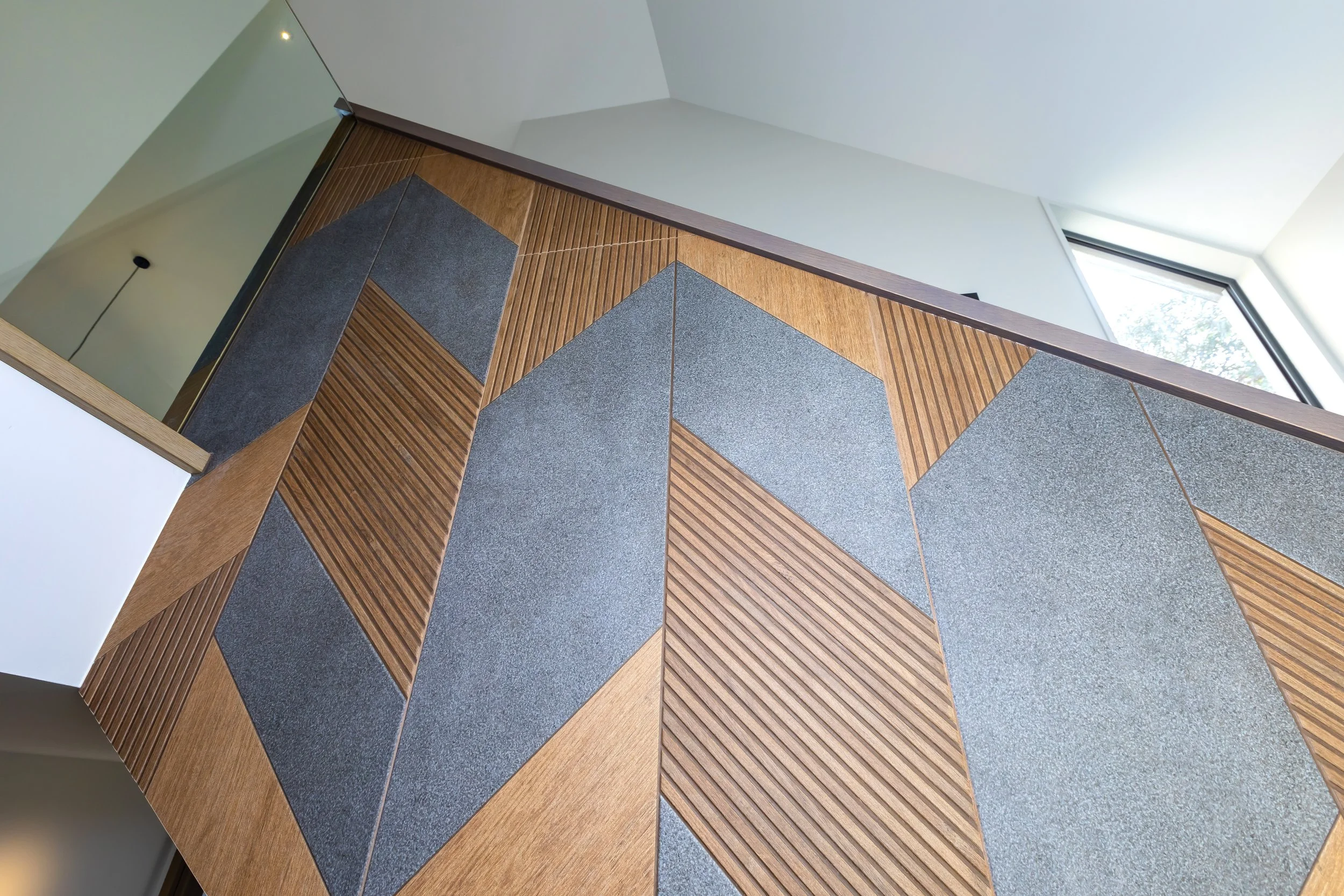 Close-up of a modern wall with wood and dark gray textured tiles arranged in a geometric pattern, a corner with a white wall, and a window letting in natural light. Stair Design by Bridget Coles Interior Design