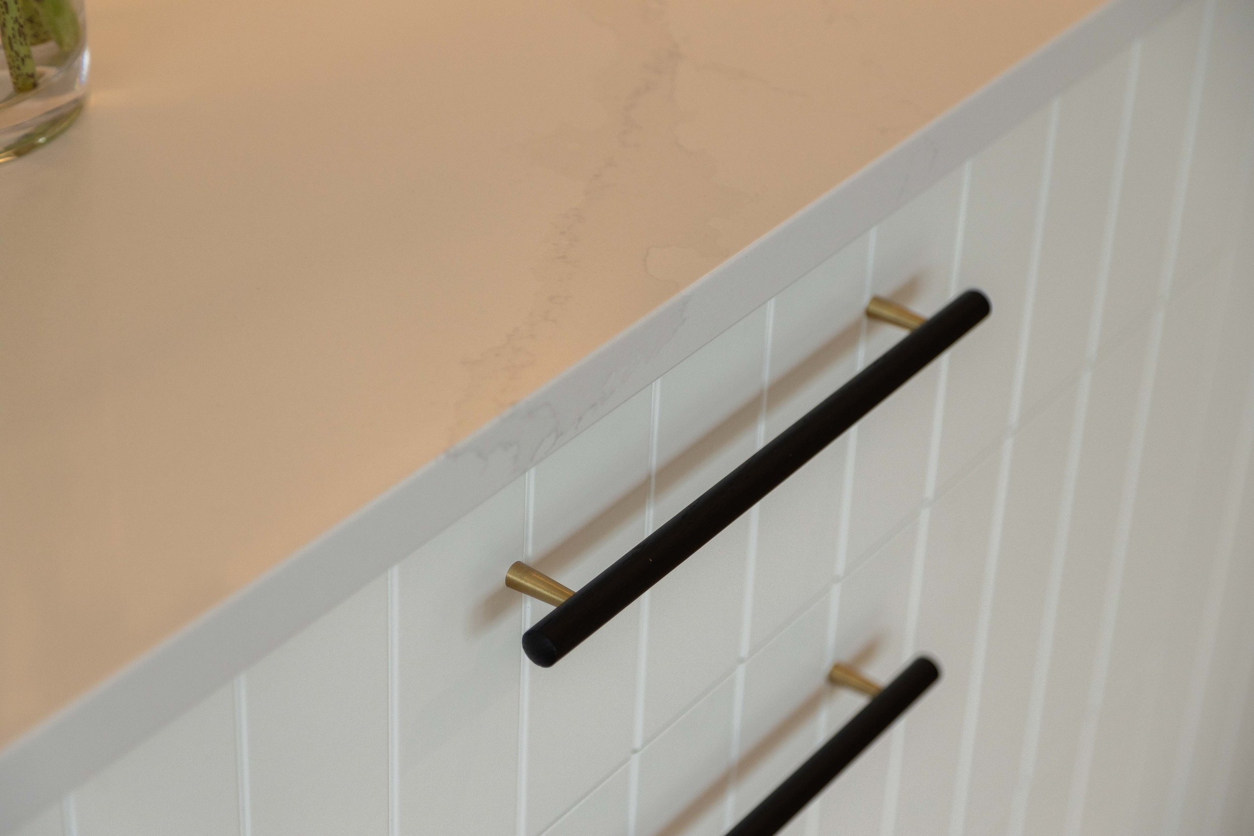 Close-up of a white tiled wall with four black and gold hooks mounted on it, and a beige counter or shelf above with a glass container partially visible in the upper left corner.