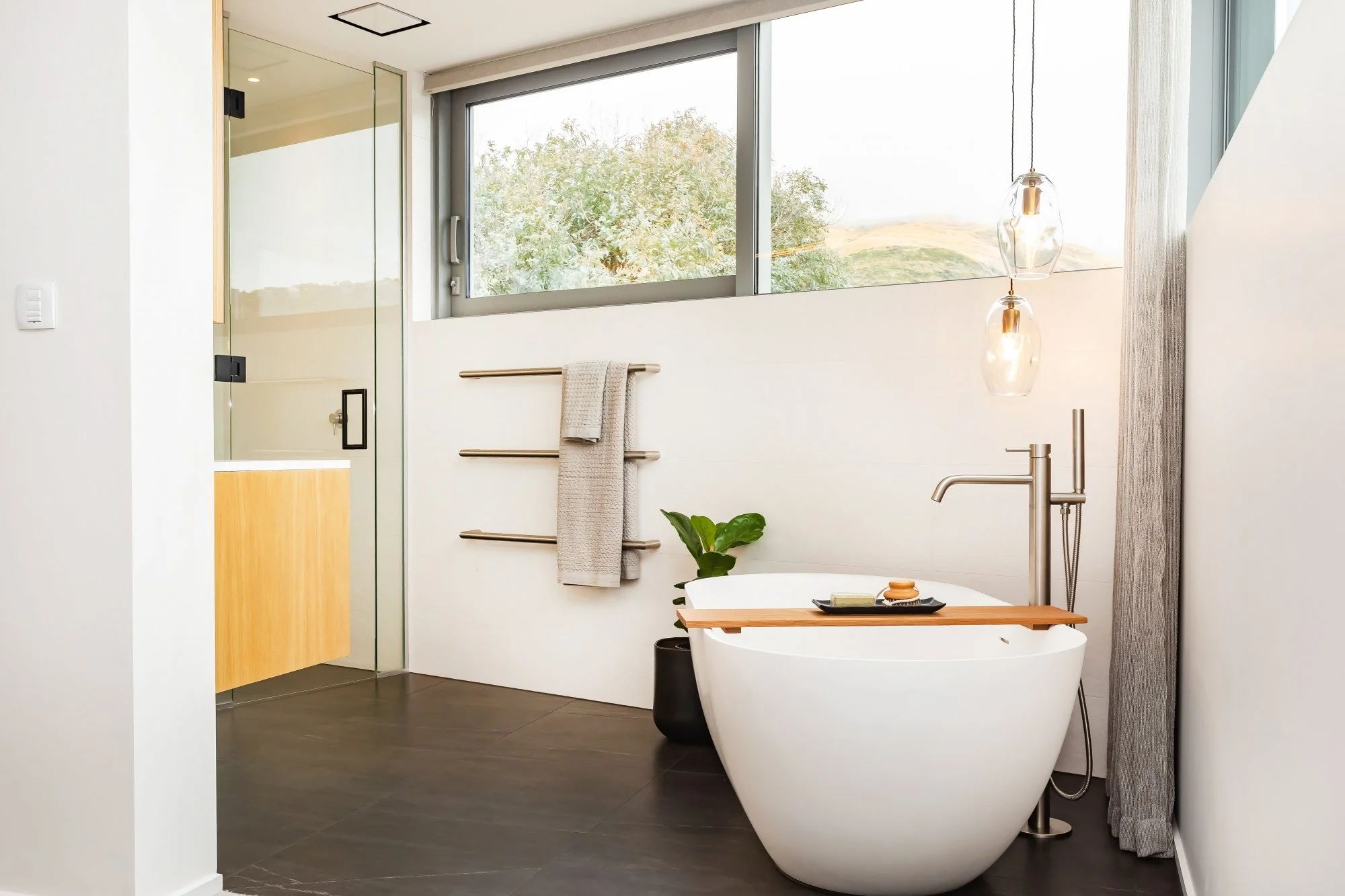 Modern bathroom with a freestanding bathtub, large window, hanging pendant lights, towel rack, small plant, and a glass-enclosed shower.