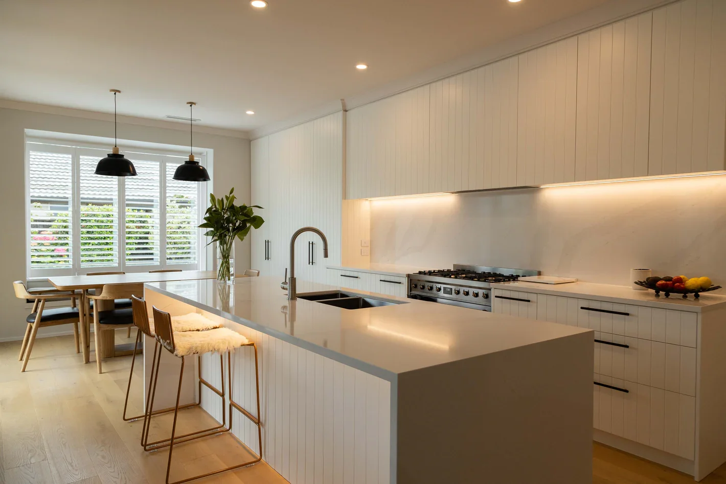 Modern kitchen with large white island, black pendant lights, white cabinetry, marble backsplash, kitchen stovetop, and windows with Norman plantation white shutters.