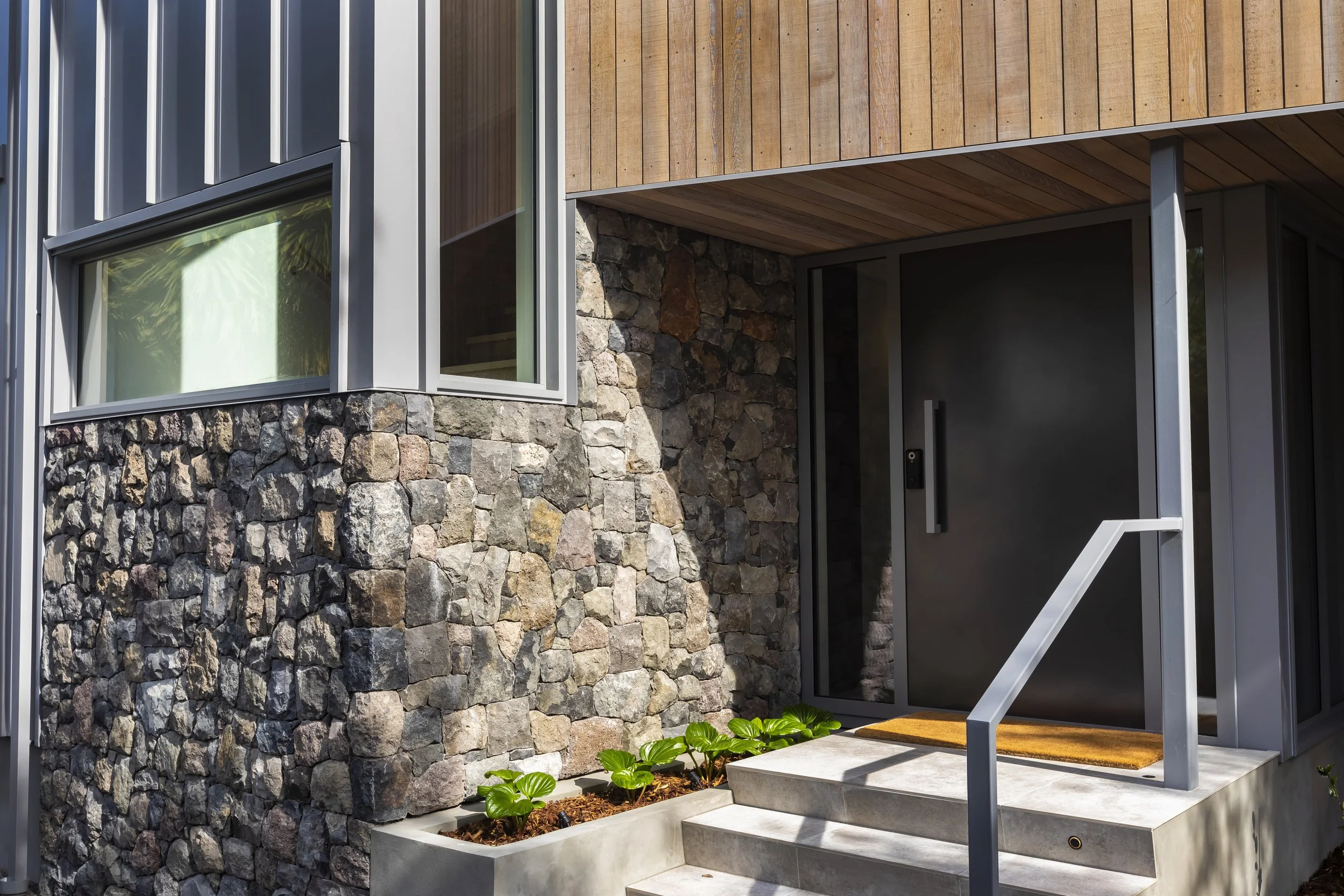Close-up of a modern house entrance with a stone wall, large window, wooden and metal siding, concrete steps, and small green plants in a planter.