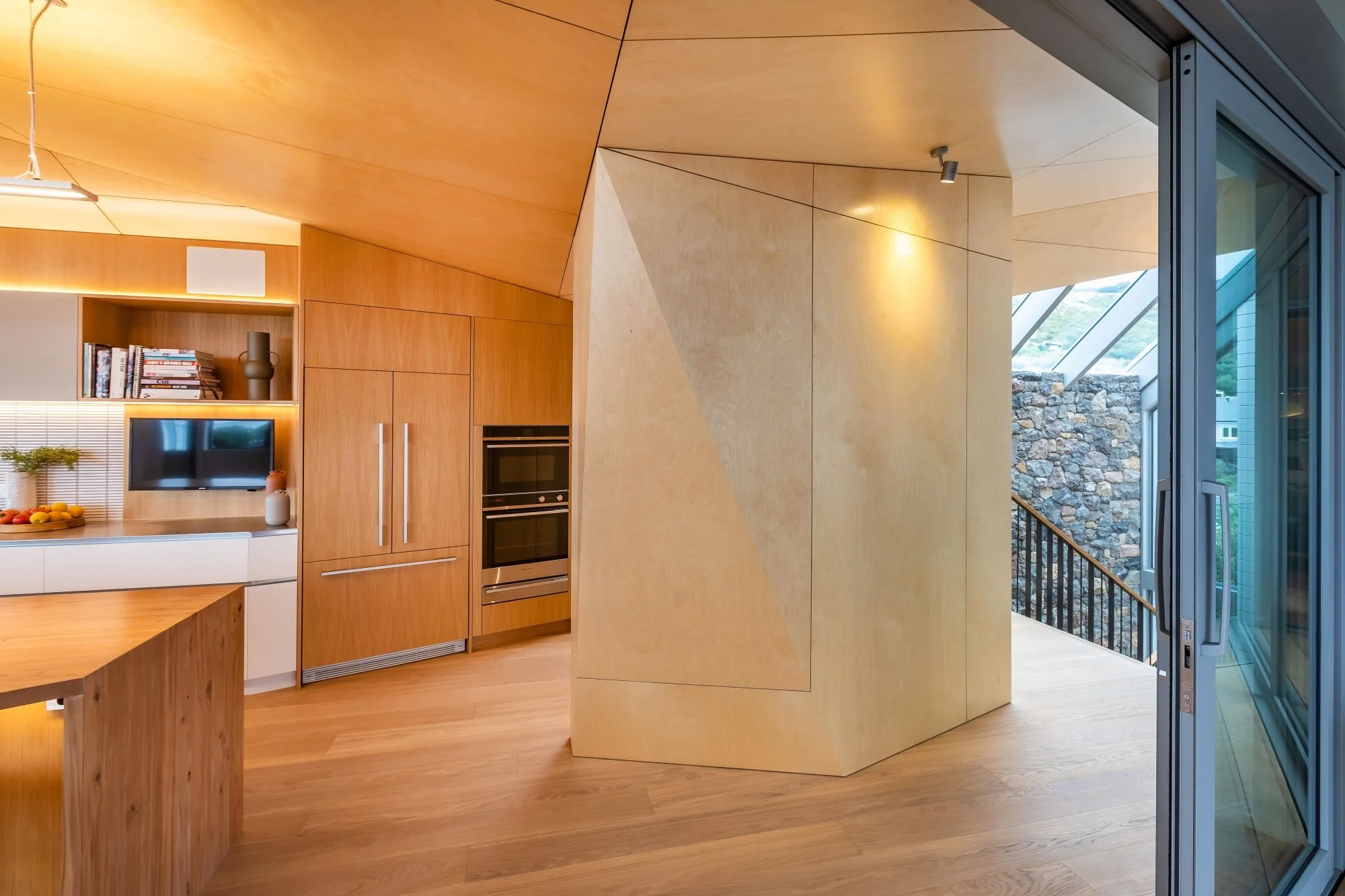 Modern kitchen and living area with light wood paneling, built-in oven, small television, books, and decorative decor. Large windows reveal a stone wall outside and allow natural light into the space.