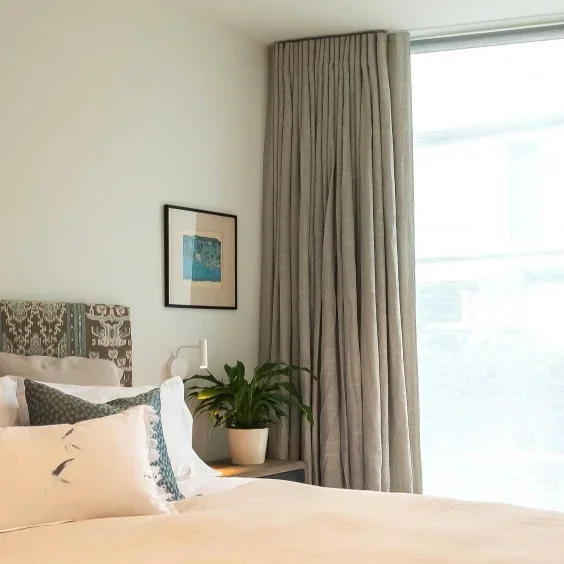 Coastal Style. A neatly made bed with white pillows and a decorative pillow, a upholstered headboard, a small lamp on a nightstand, a potted plant, framed artwork on the wall, and beige blackout curtains beside a large window in a bright bedroom.