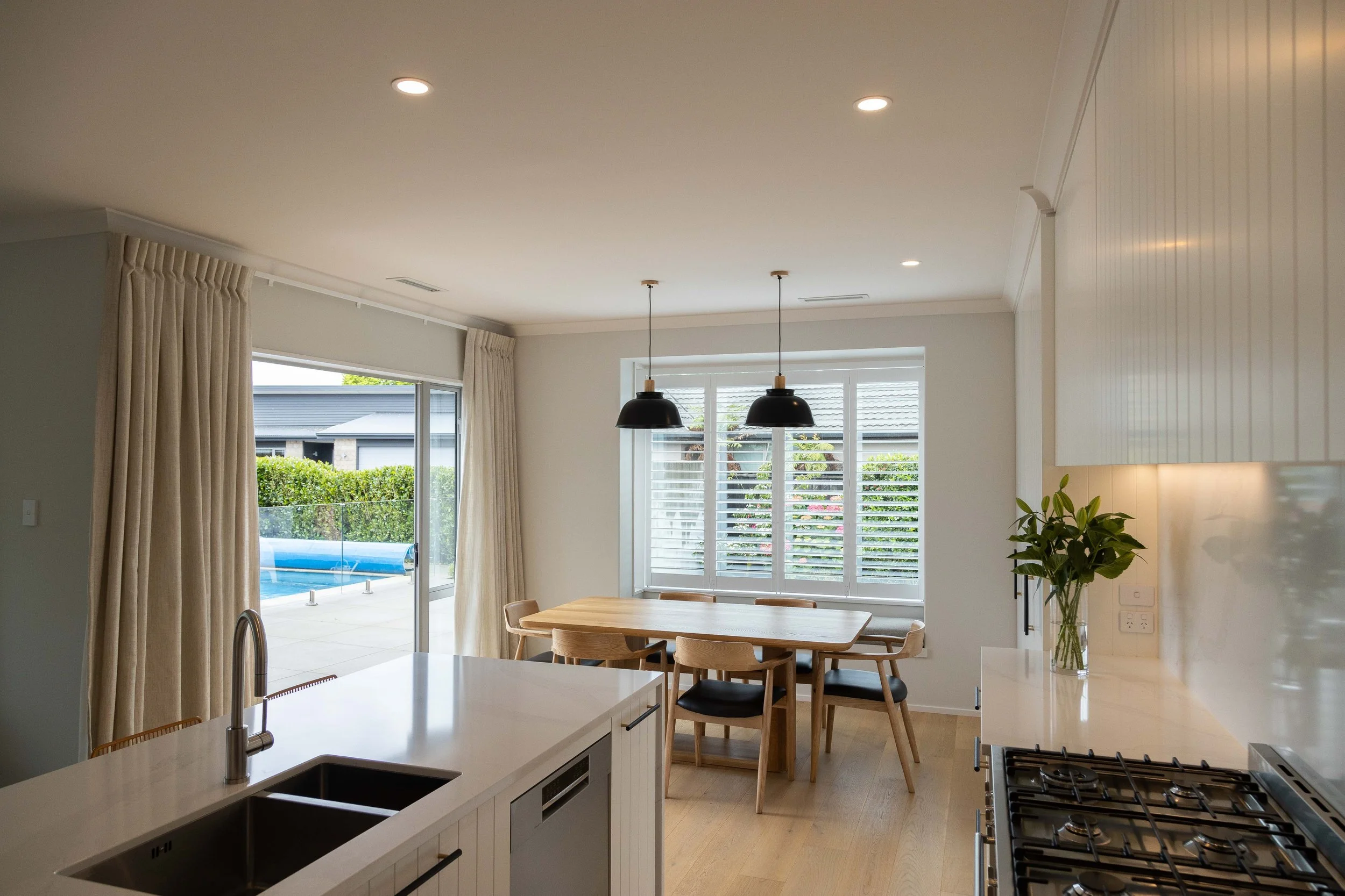 Modern kitchen and dining area with large window, sliding glass door, pendant lights, wooden dining table with chairs, potted plant, and view of backyard with pool.