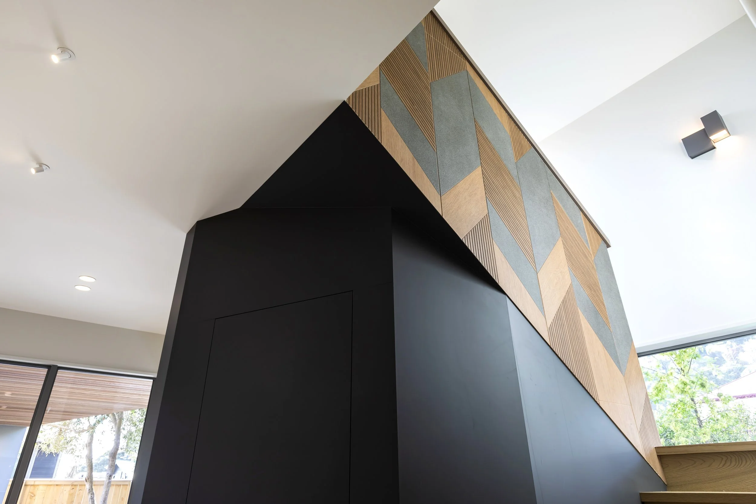 Interior of a modern home or building featuring a ceiling with recessed lights, a wall with geometric wood and gray paneling, and large windows showing trees outside. Stair Design by Bridget Coles Interior Design
