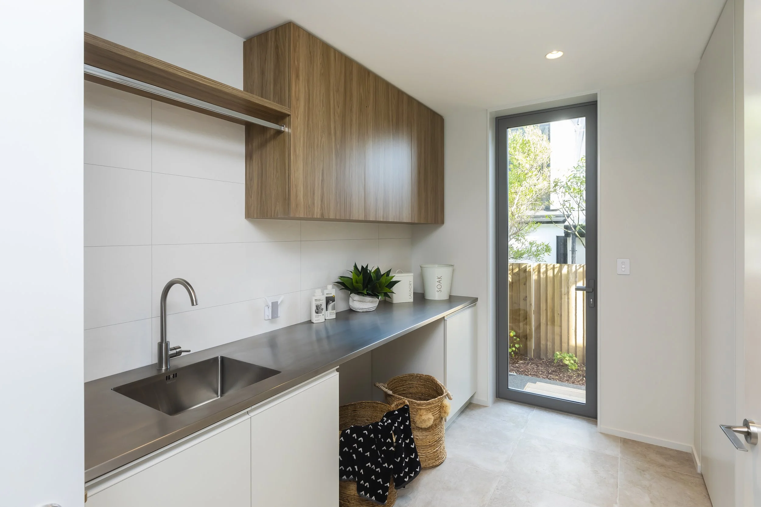 A modern laundry room featuring white cabinets, a stainless steel sink, a countertop with laundry essentials, a potted plant, and a glass door leading outside with greenery visible. Laundry Design by Bridget Coles Interior Design