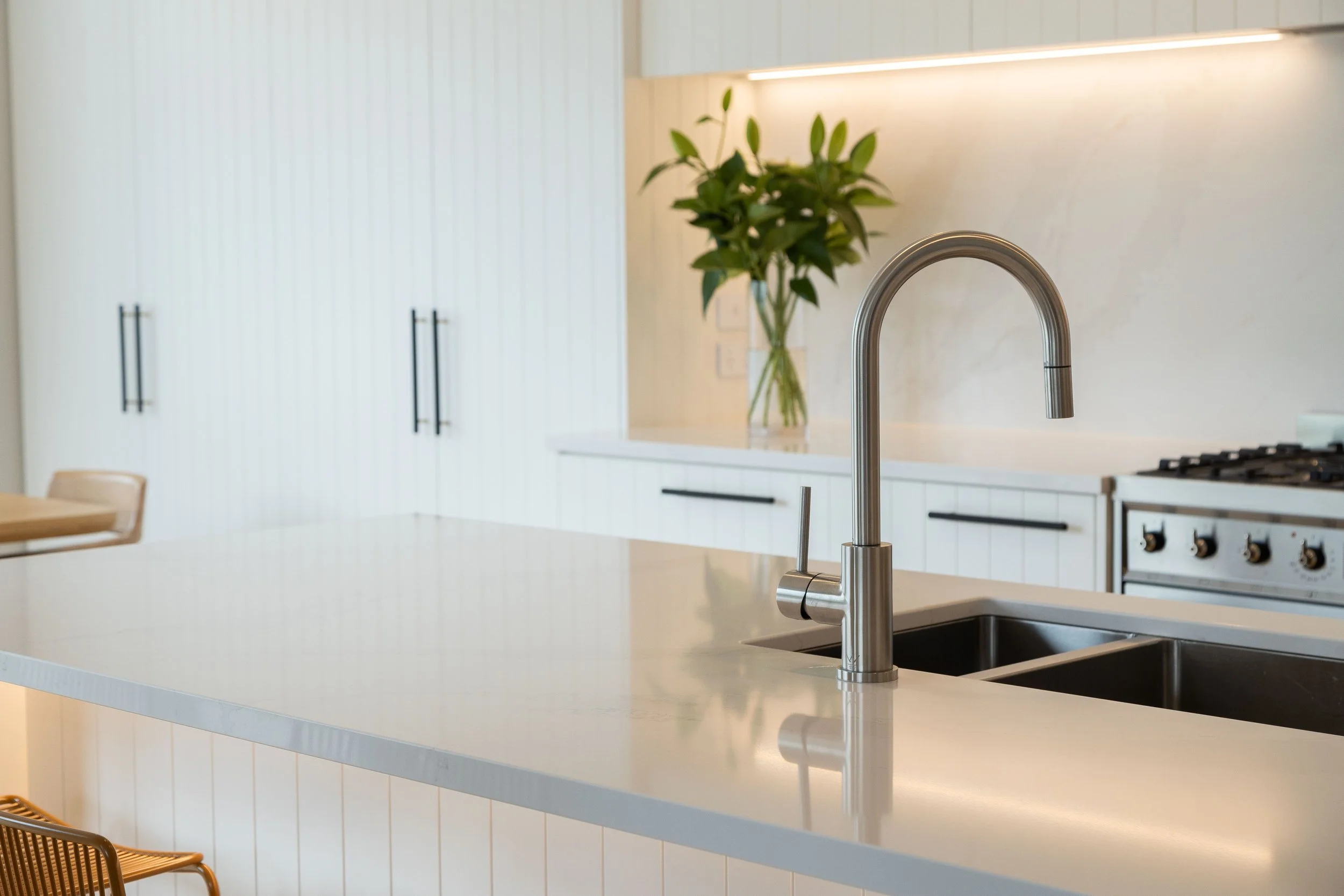 Modern kitchen with a white countertop and a stainless steel faucet, a vase with green leaves on the counter, white cabinets with black handles, and a stove in the background.