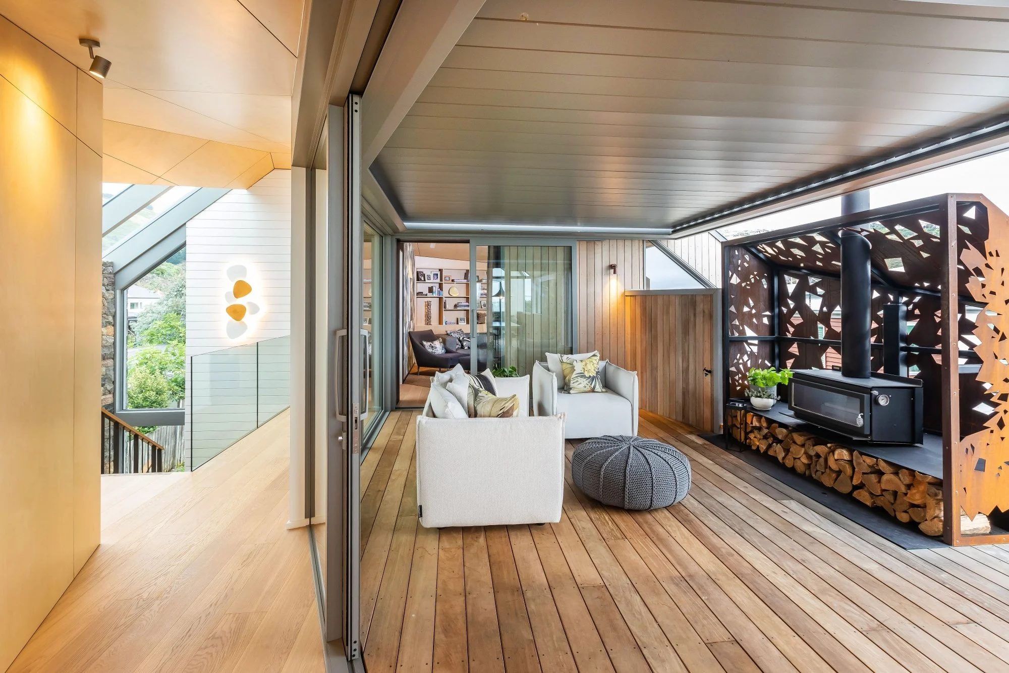 Interior view of a modern living space with wooden floors and ceiling, glass sliding doors, and a cozy seating area with white and patterned cushions, a black stove on a wood storage area, decorative wooden panels, and a shelf with books.