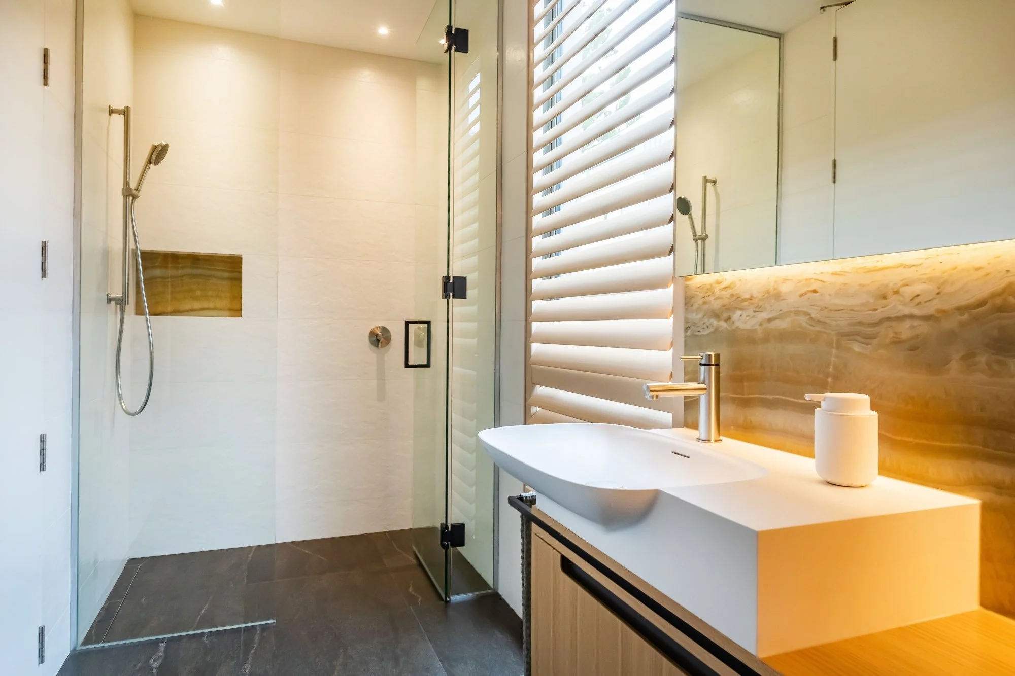 Modern bathroom with a glass shower enclosure, white sink with a brass faucet, a decorative onyx backsplash, a large mirror, and window shutters.