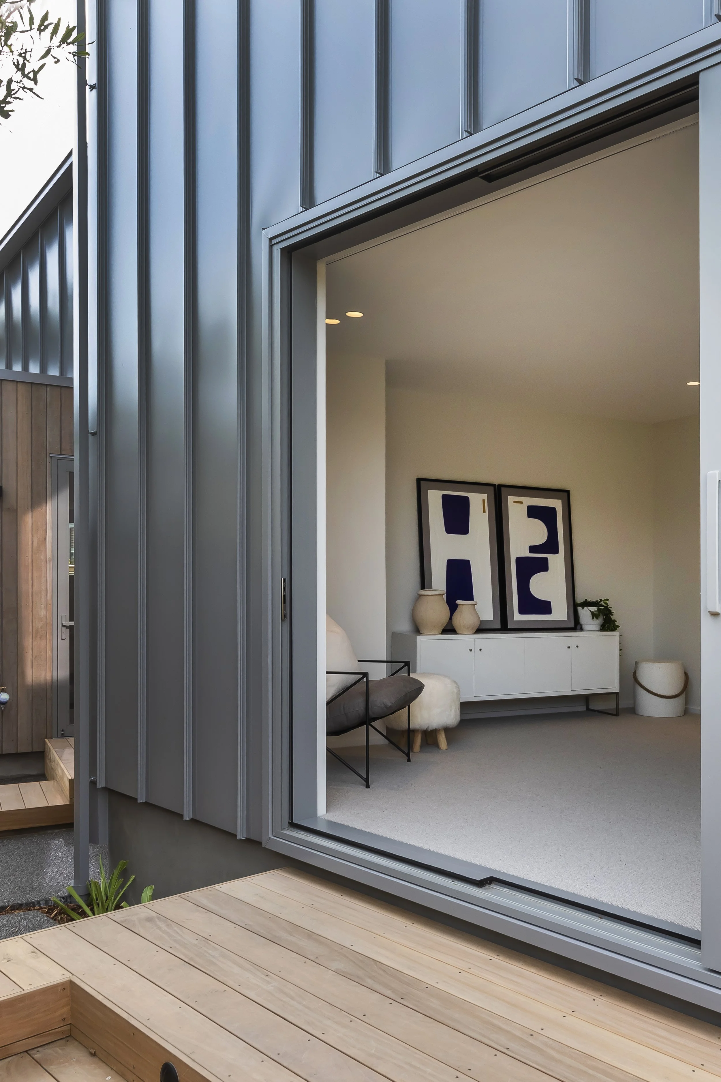 A view of a modern house exterior with a large open window showing a minimalist interior with framed artwork, vases, a small plant, and modern furniture. The house features gray metal siding and wooden adjacent steps or deck.