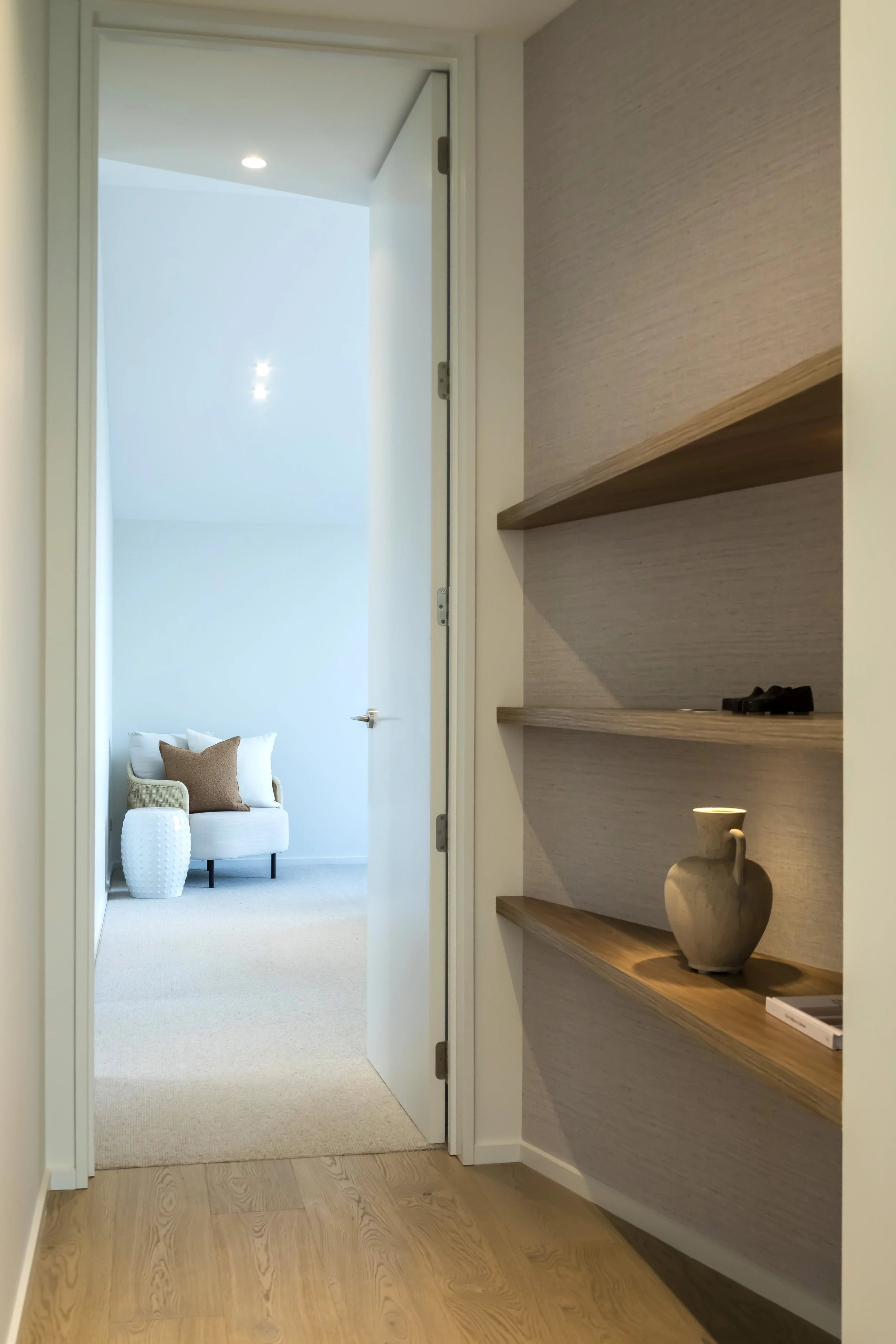 A view through a doorway into a bright room with a white armchair, a pillow, a white textured vase, and a small side table. The room has light-colored walls and a high ceiling with skylights.