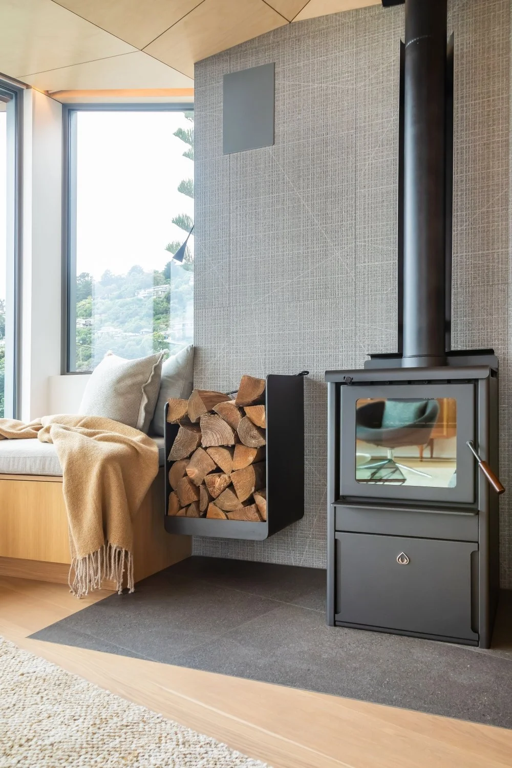 A cozy indoor corner with a wood-burning stove, a pile of firewood, a beige sofa with cushions and a blanket, and large windows showing outdoor greenery.