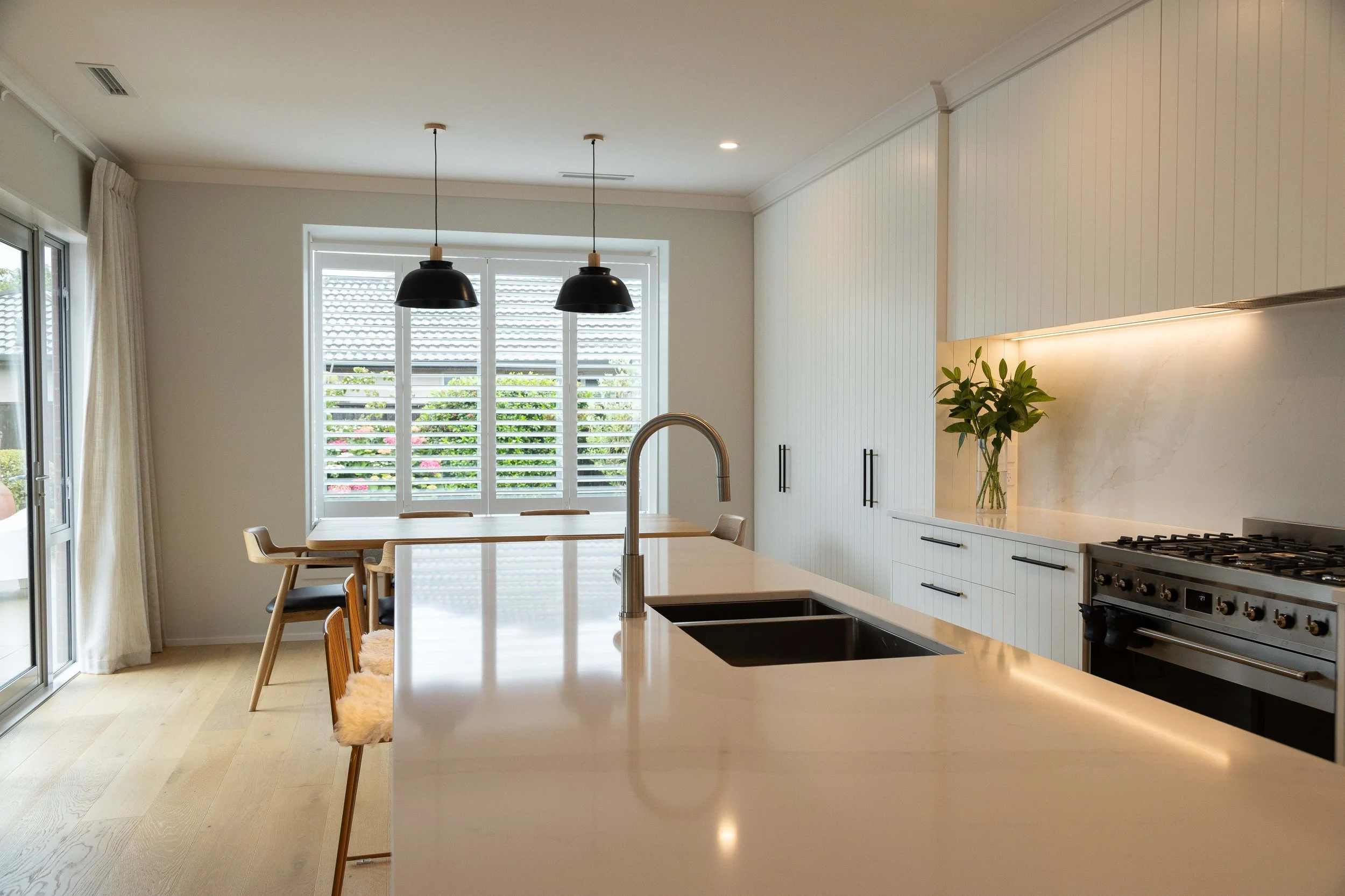 A modern kitchen with white cabinetry, a beige countertop, a black stove, a sink with a faucet, and a vase with green leaves on the counter. There are large windows with plantation shutters and pendant lights hanging over a dining table.