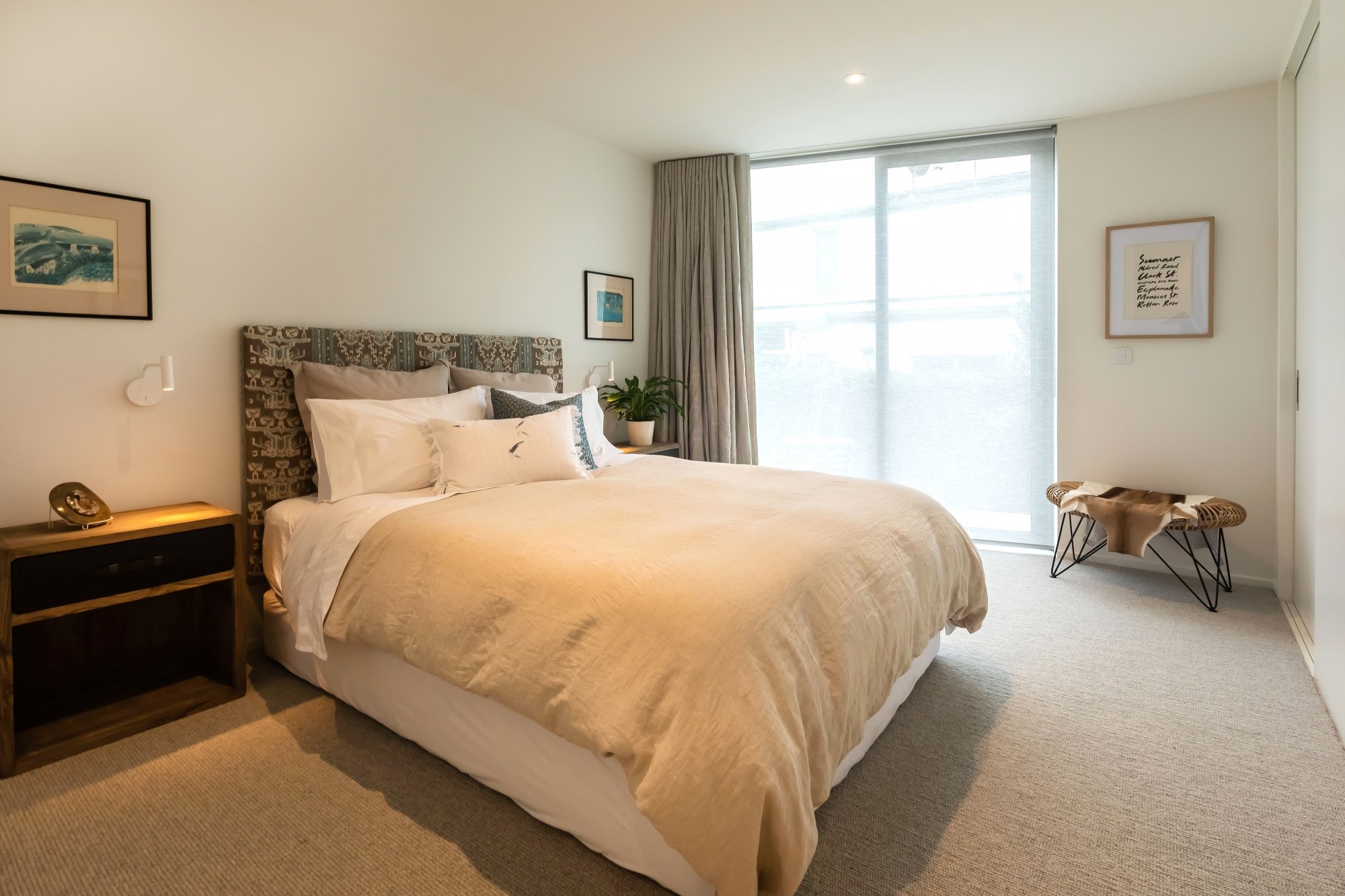 A cozy bedroom with a large bed covered in beige bedding and multiple pillows. There is a dark wooden nightstand with a vintage-style alarm clock and a telephone. The headboard is upholstered with patterned fabric. The wall features framed artwork. A