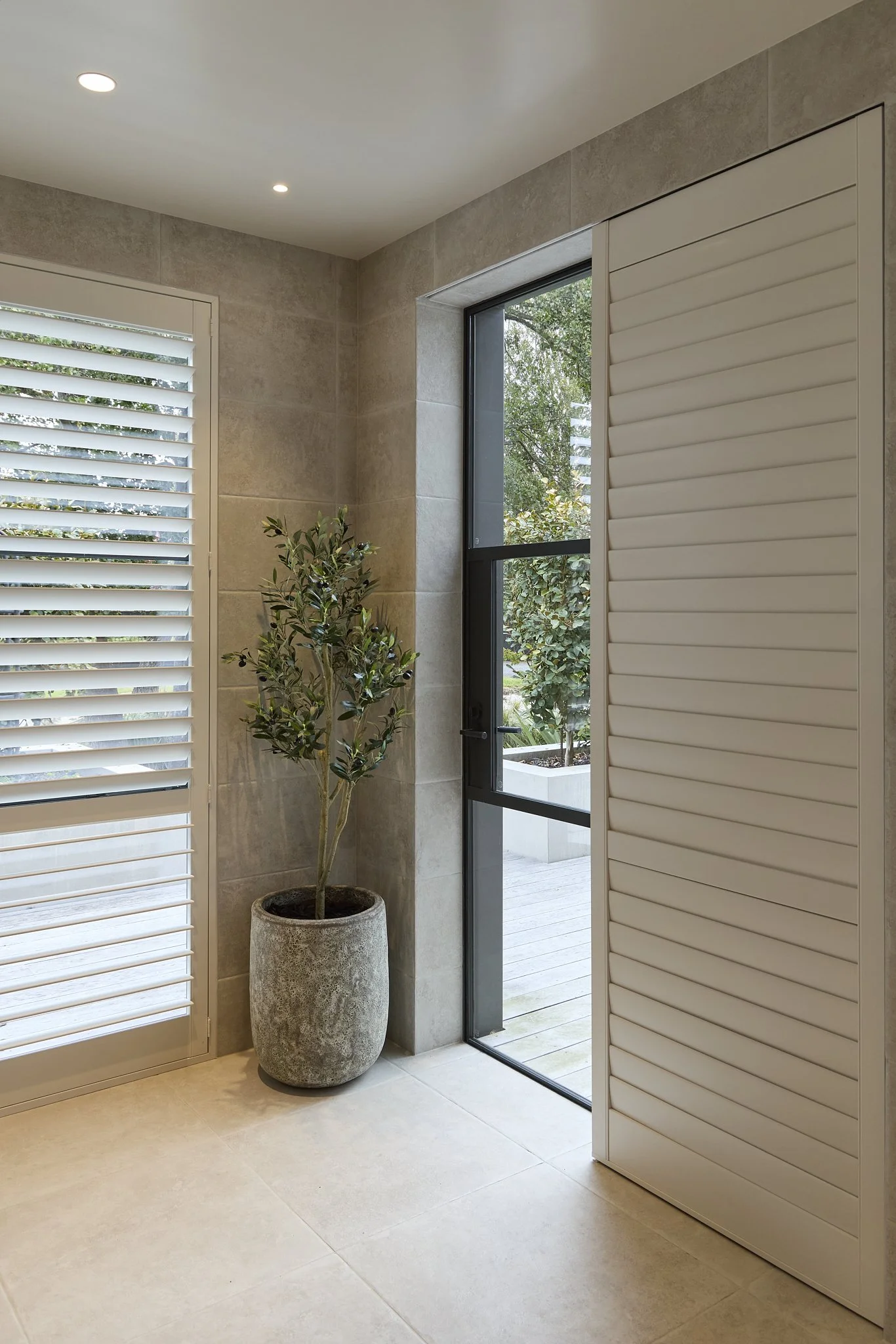 Indoor space with beige tiled floor and walls, a tall potted plant next to a window, white louvered shutters on the left, a sliding glass door leading outside, and a white sliding shutter by Norman. cleverly intergrated into the tiling .