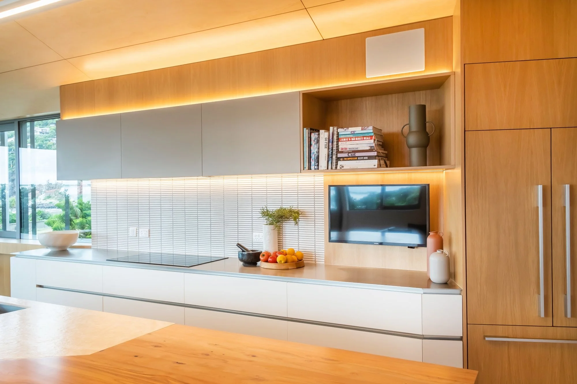 Modern kitchen with white countertop, wooden cabinetry, and a wall-mounted TV. Decor includes vases, and a bowl of fresh fruit on the counter. Large windows show greenery outsideInterior design Bridget Coles Interior Design Christchurch BCSTUDIO