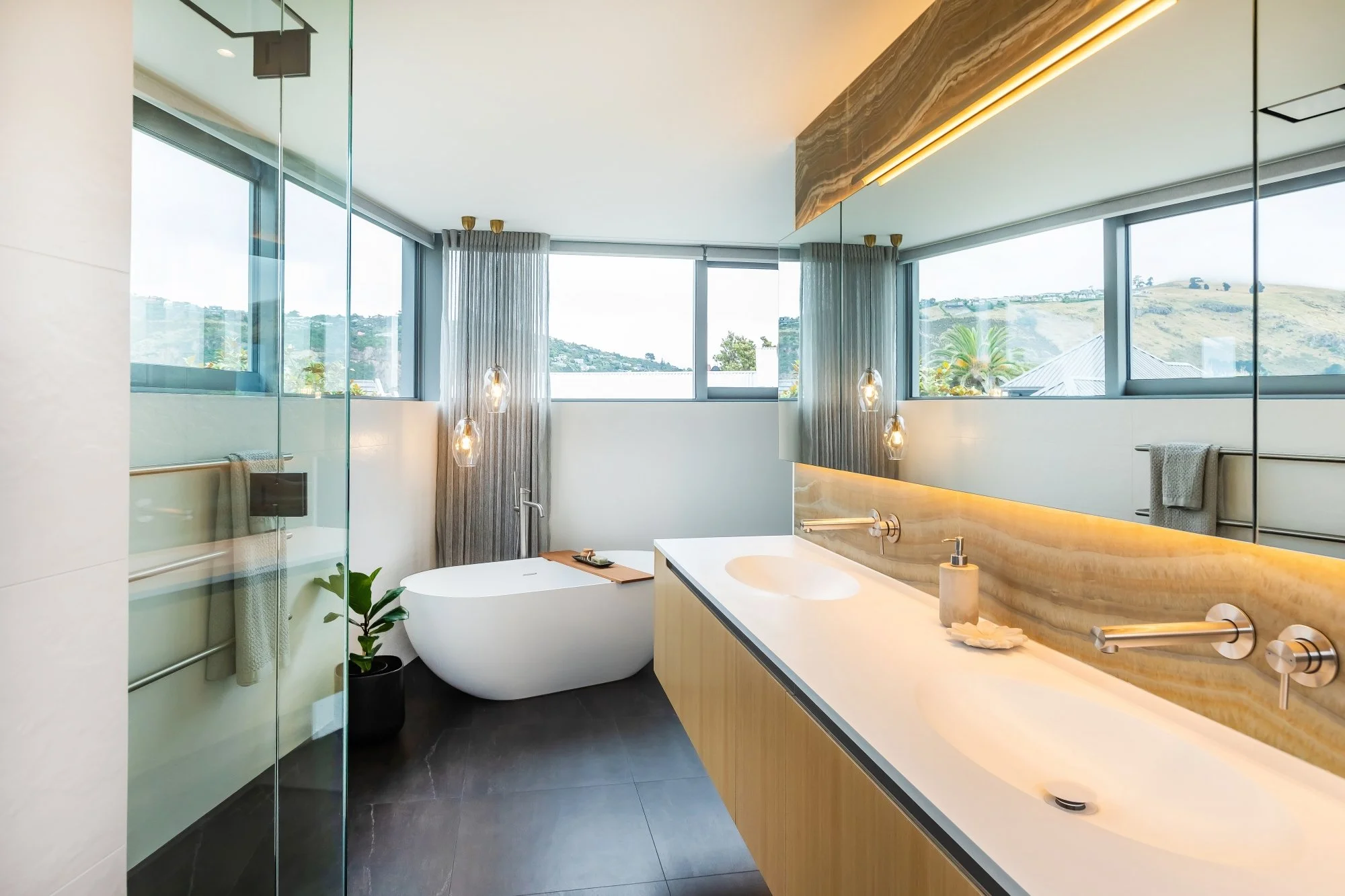Modern bathroom with large windows showing a hillside view, a freestanding bathtub, a wooden vanity with a white sink, hanging pendant lights, and a plant near the shower area.