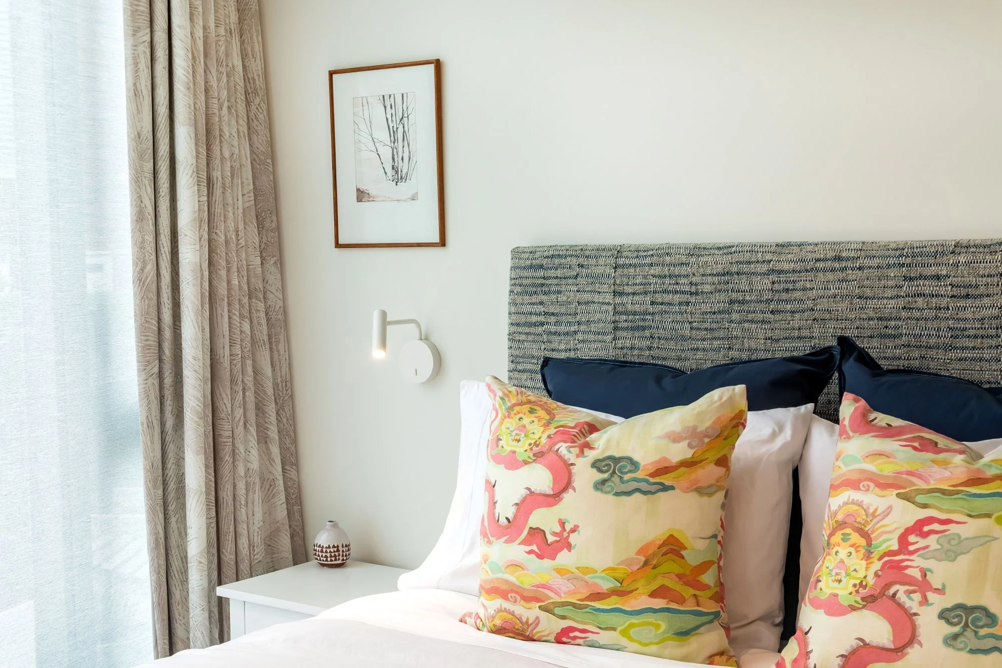A hotel room bedroom corner with a bed featuring colorful dragon-themed pillows, a white bedside table with a decorative lamp, beige patterned curtains, a wall-mounted reading light, and a framed black-and-white line drawing of trees on the wall.