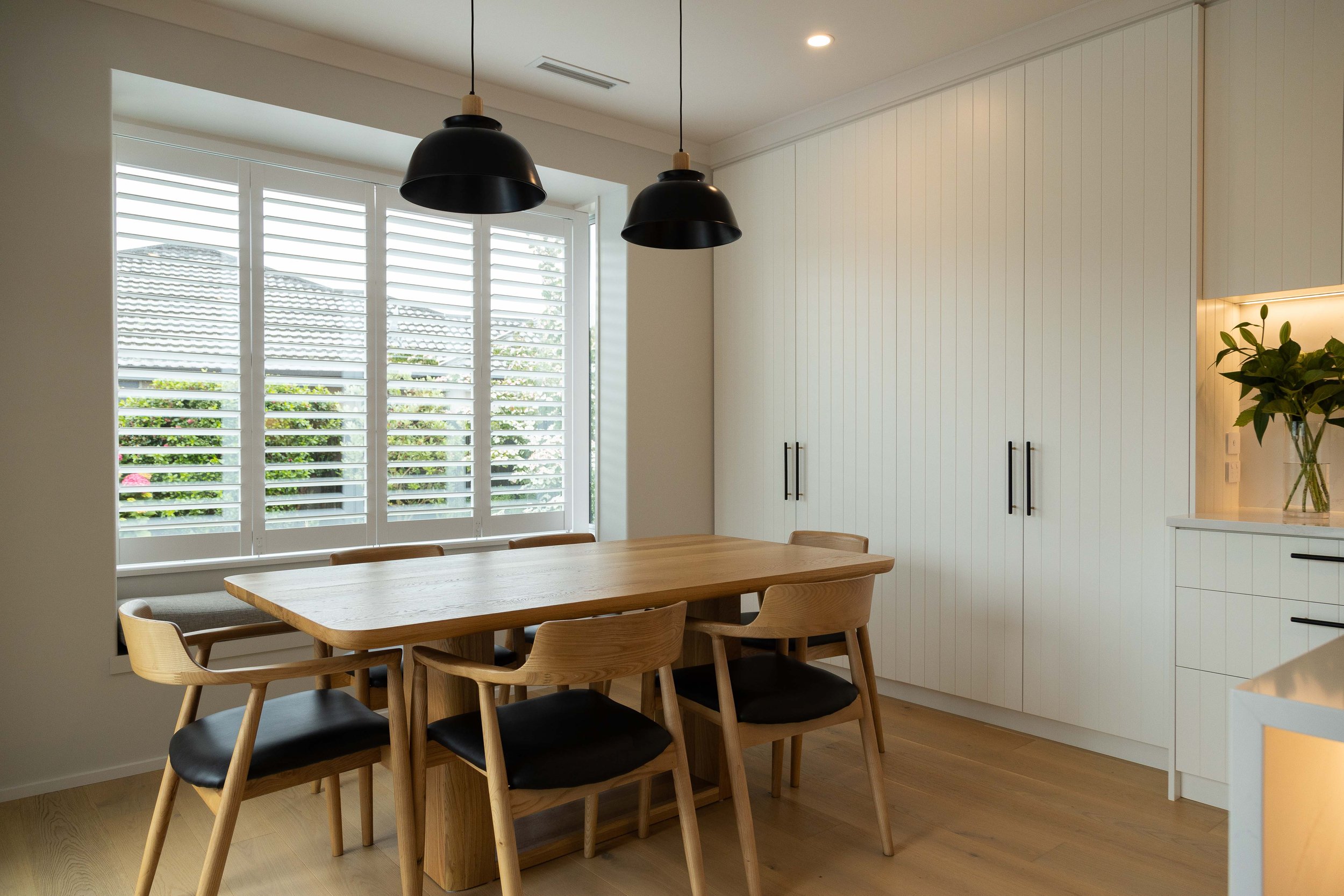 A modern kitchen with a wooden dining table surrounded by wooden chairs with black seats, large window with white Norman shutters, white cabinets, hanging black pendant lights, and a potted plant on a counter.