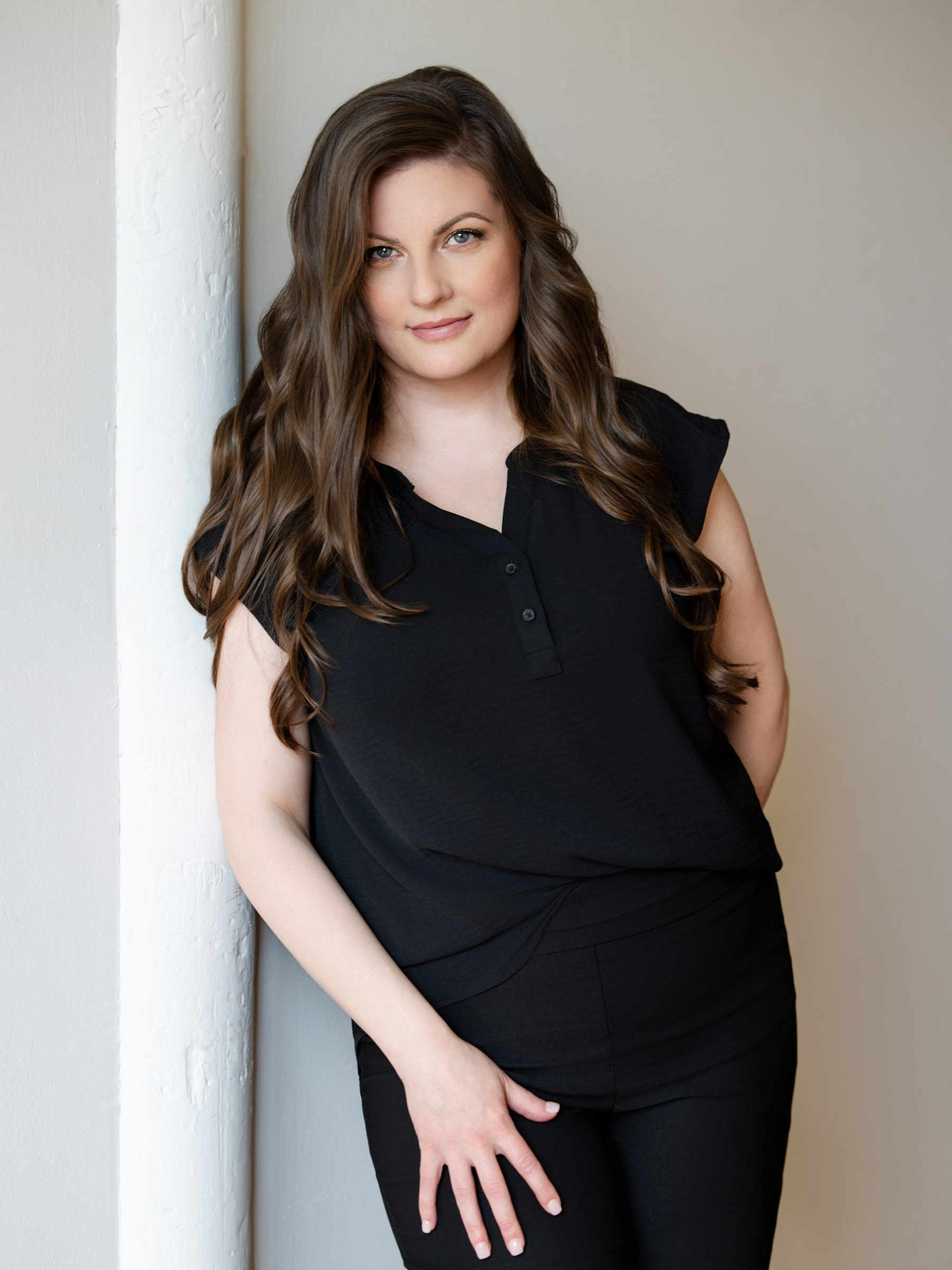 A woman with long wavy brown hair and blue eyes is leaning against a white wall. She is wearing a black short-sleeved top and matching black pants, looking at the camera with a subtle smile.