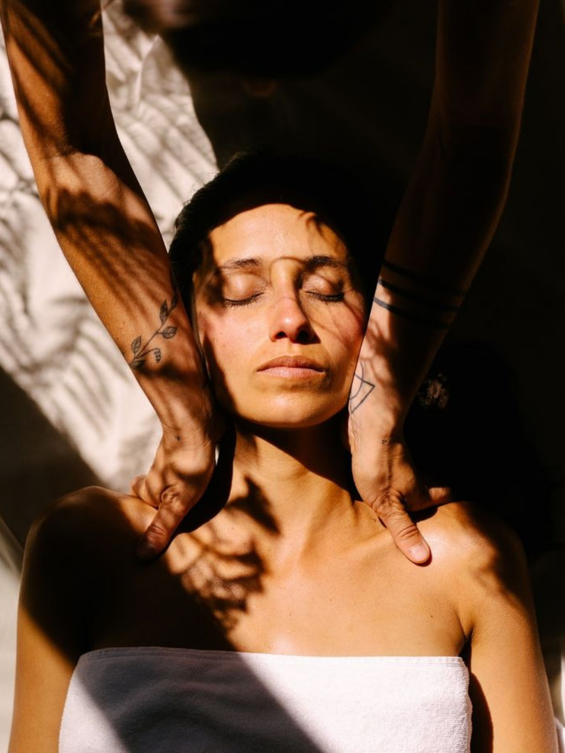 A woman with closed eyes receives a massage, lying on a bed, with shadows of leaves cast on her face and body.
