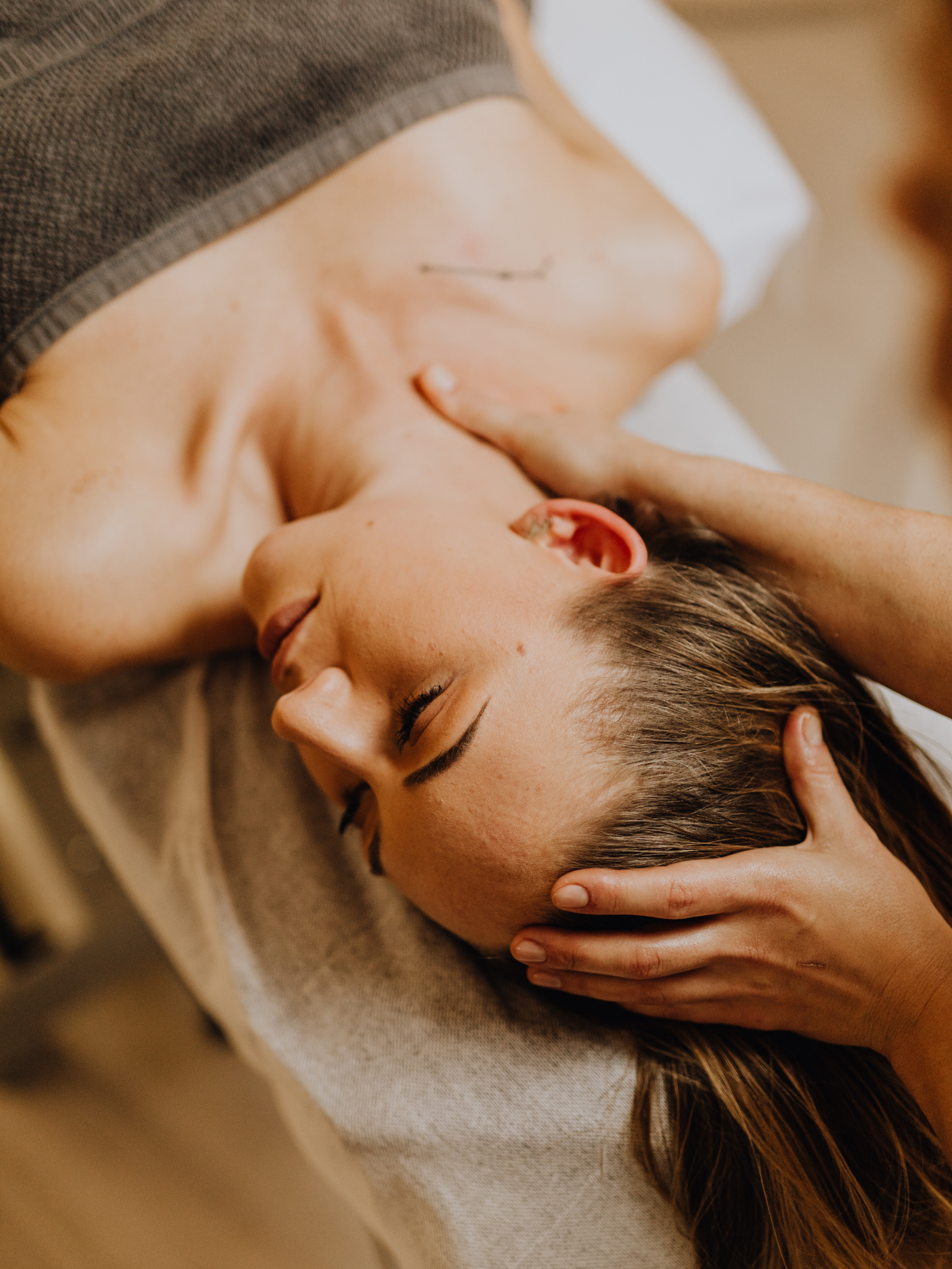A woman receiving a massage on her head and neck, lying on her back with eyes closed.