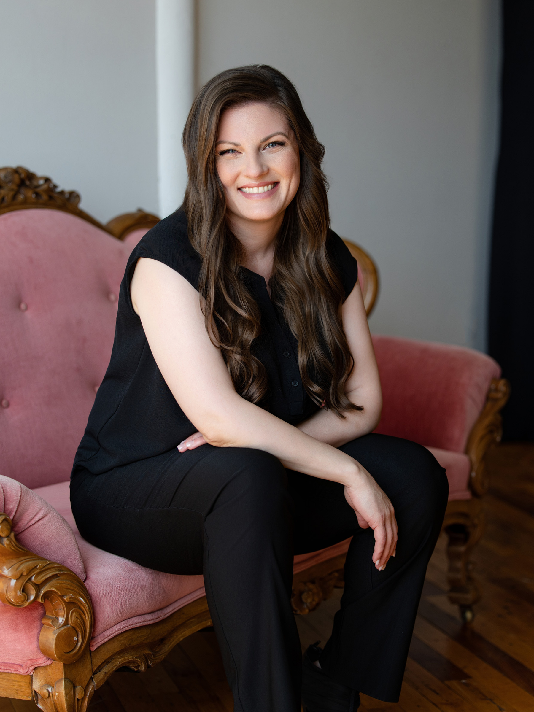 A woman with long wavy brown hair sitting on a vintage pink upholstered sofa with carved wooden details, smiling at the camera.