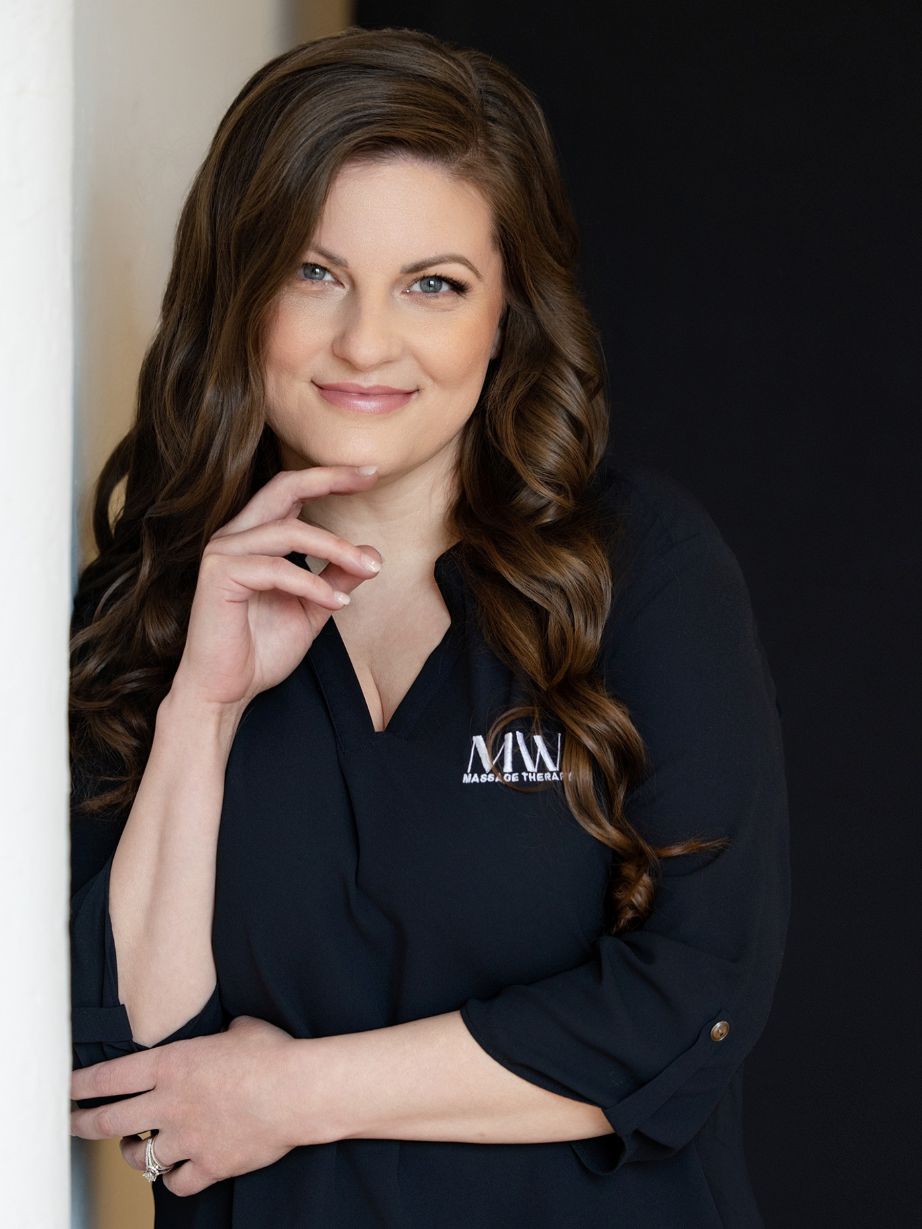 A woman with long wavy brown hair and blue eyes, smiling softly, wearing a black top with a logo on it, posing with her hand near her chin.