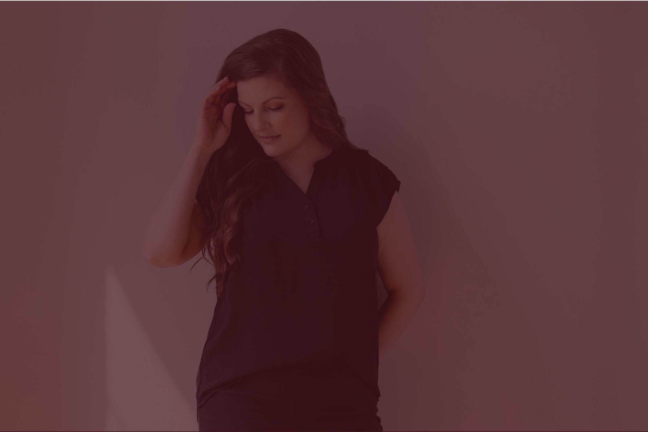A woman with long, wavy brown hair wearing a black top, standing against a plain background, looking down with her hand touching her forehead.