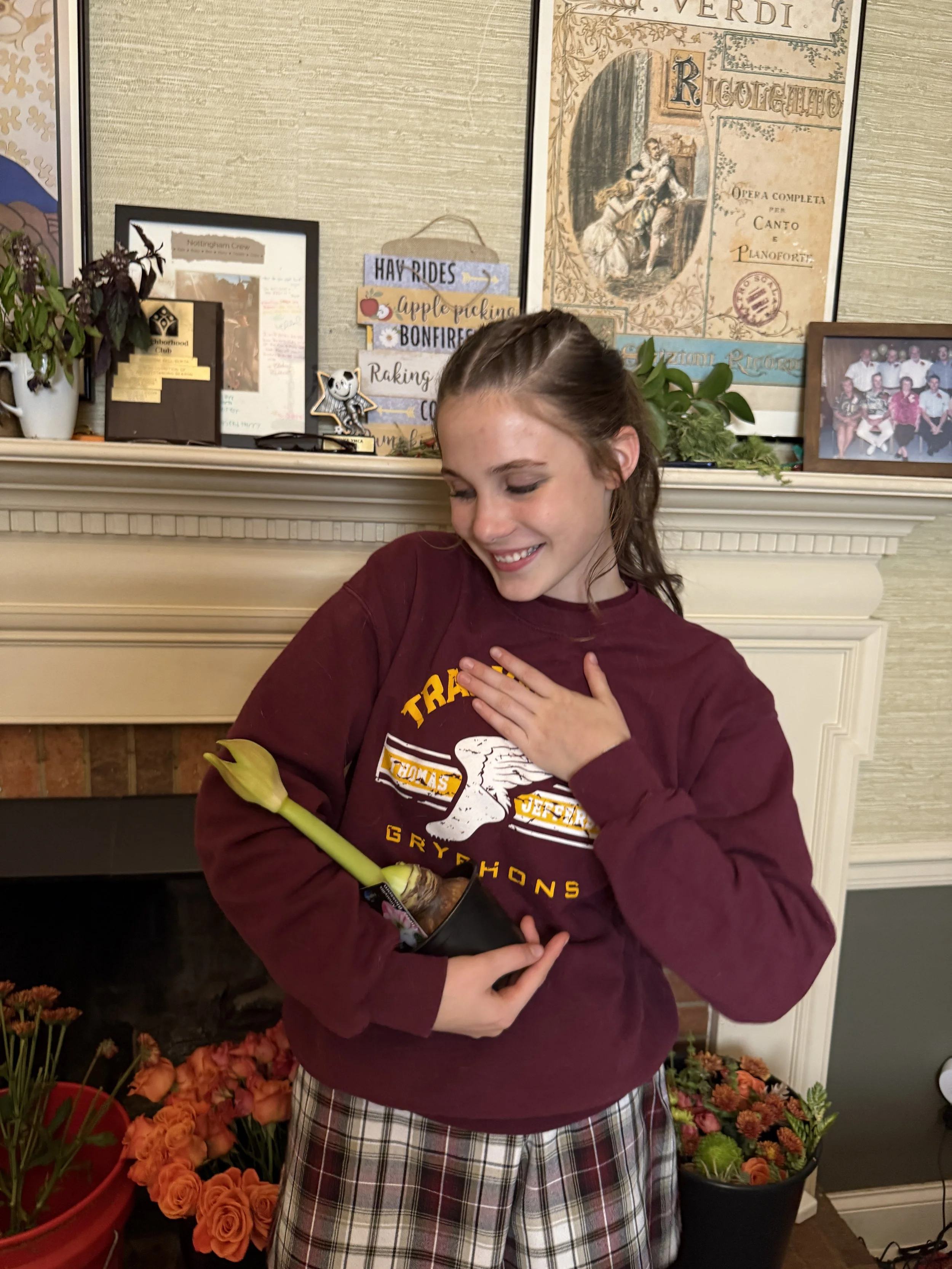 A girl in a Gryffindor sweatshirt holding a potted flower and smiling indoors near a fireplace with flowers and framed pictures behind her.