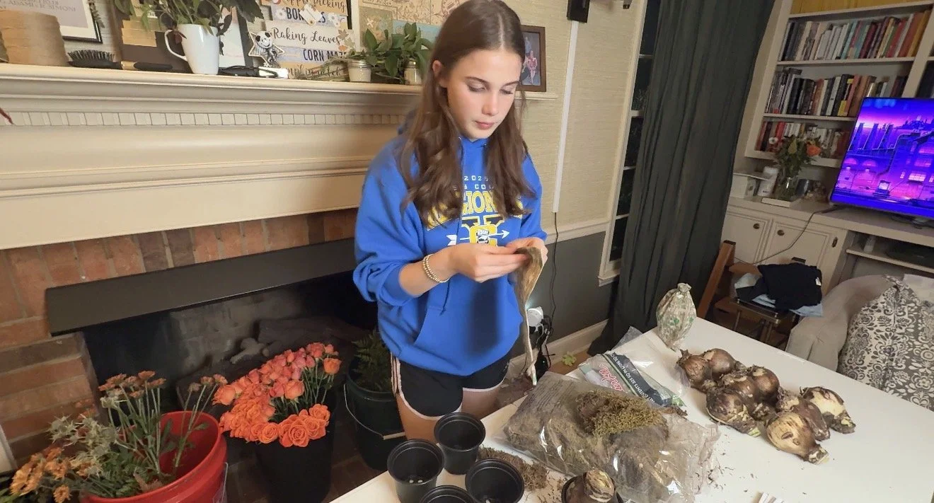 A young woman wearing a blue hoodie and black shorts stands at a table, handling some plants or soil. There are pots, a plant in a plastic bag, and several large bulbs or roots on the table. The background shows a living room with a fireplace, booksh