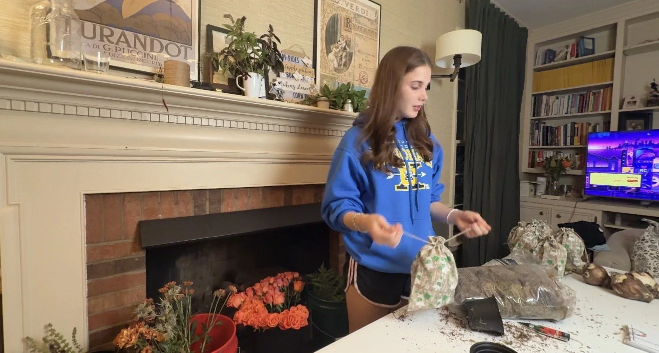 A young woman with long brown hair wearing a blue hoodie and black shorts, standing at a table with several small gift bags, with a fireplace, bookshelves, and a TV in the background.