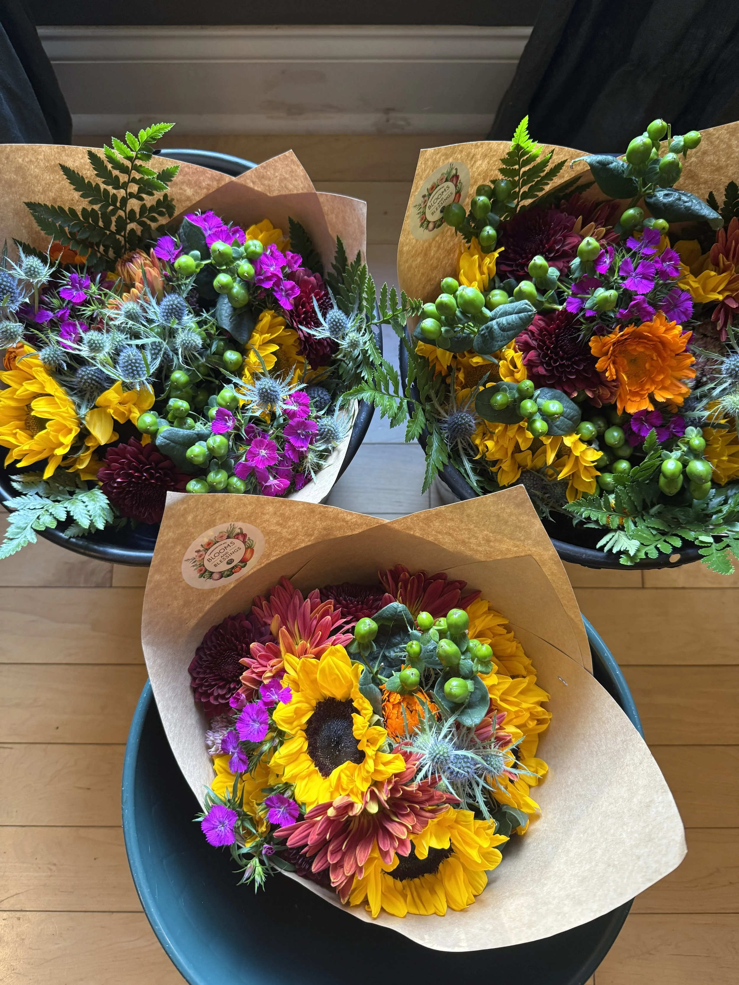 Three vibrant bouquets of flowers with sunflowers, purple and maroon blooms, green berries, and fern leaves, wrapped in brown paper.