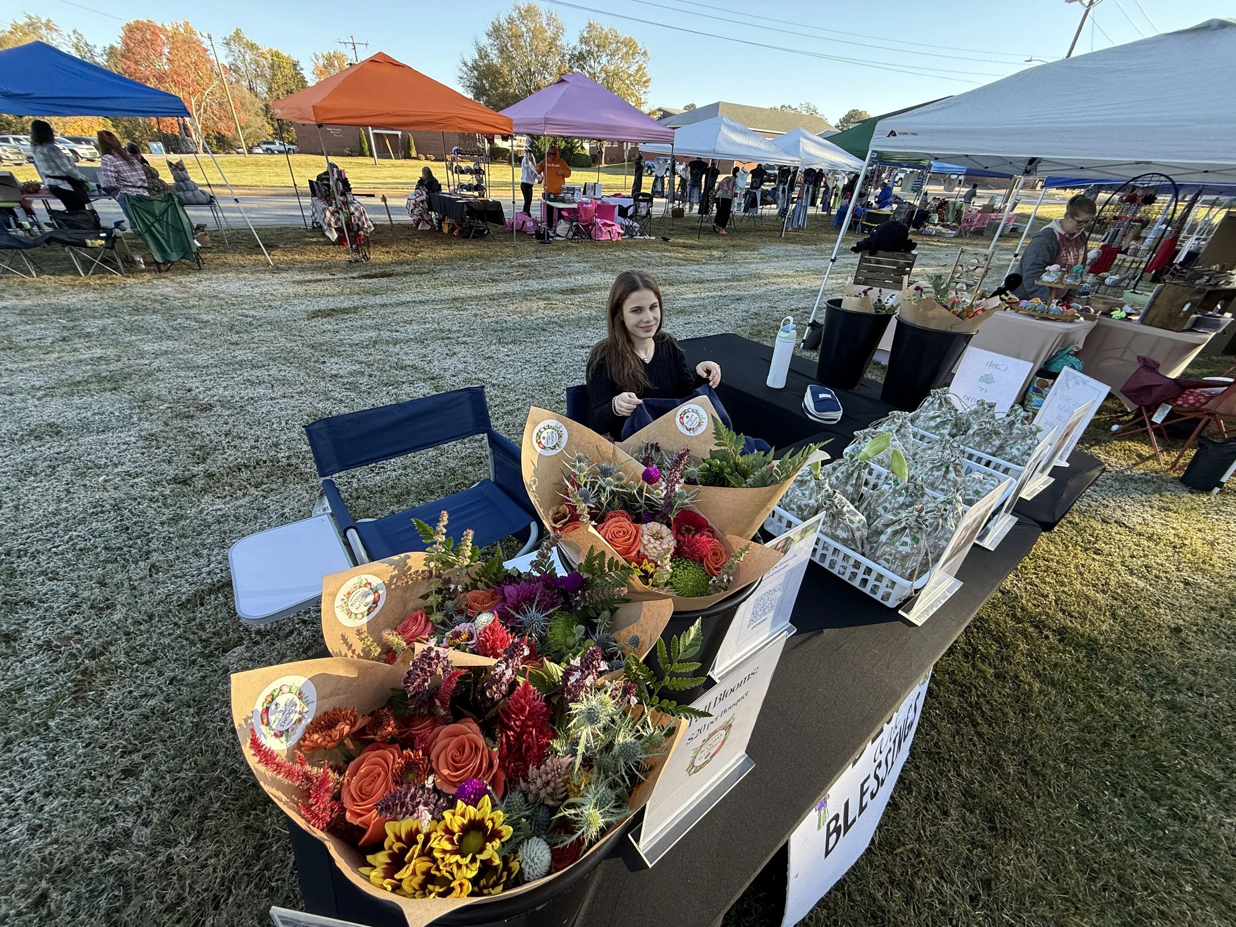 A woman sitting at a table of flower bouquets at an outdoor market during daytime, with tents and vendors in the background.