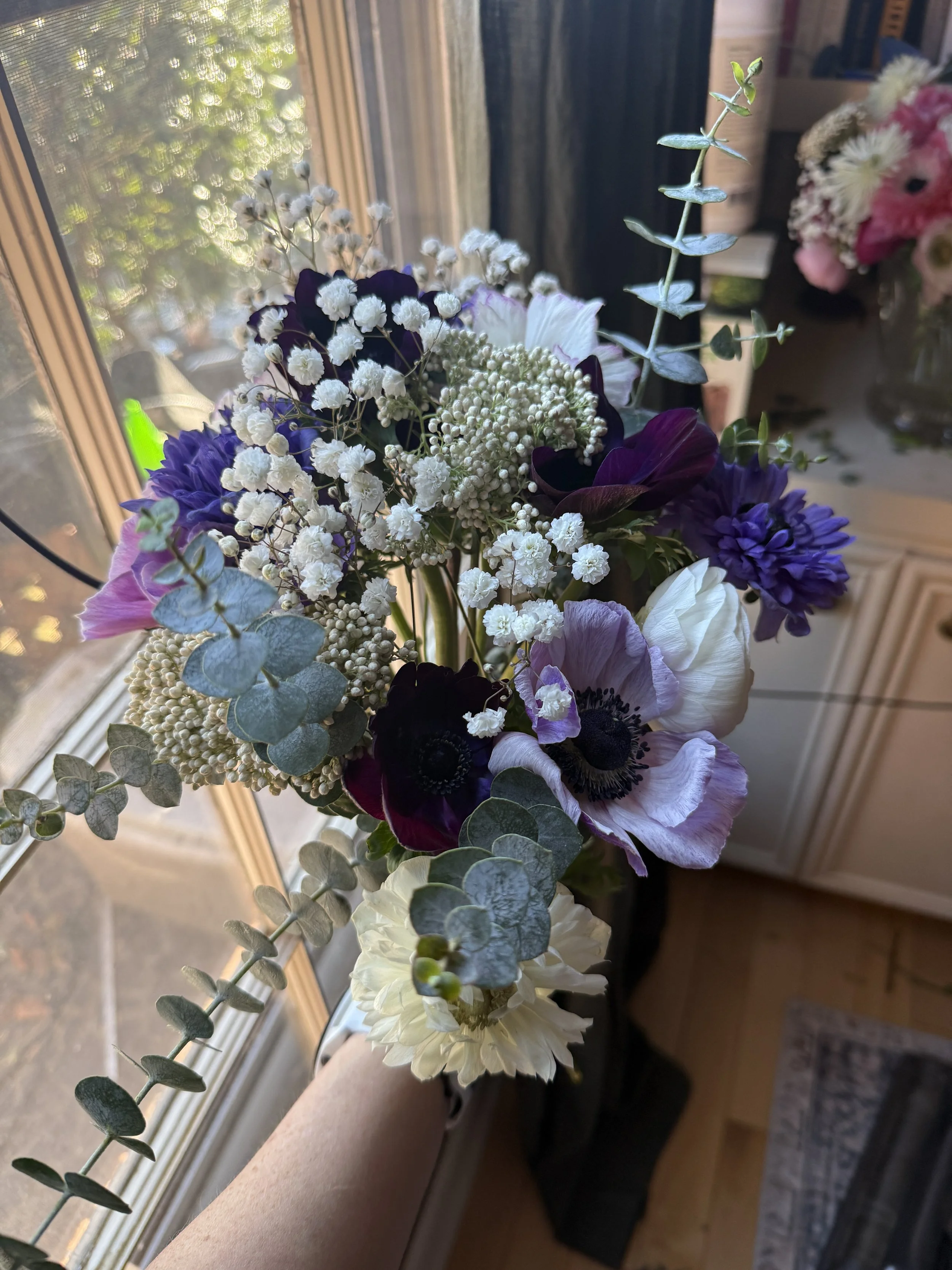 A hand holding a mixed bouquet of purple, white, and dark red flowers with greenery, near a window with sunlight coming through.