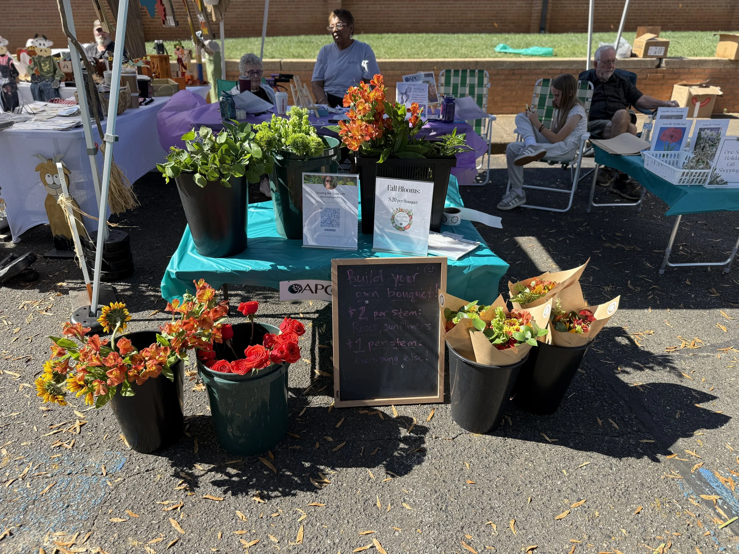 A flower stand at an outdoor market featuring vases of flowers including roses and sunflowers, with signs advertising bouquets for $20 and build-your-own options, surrounded by tables with books and crafts, and people sitting in the background.