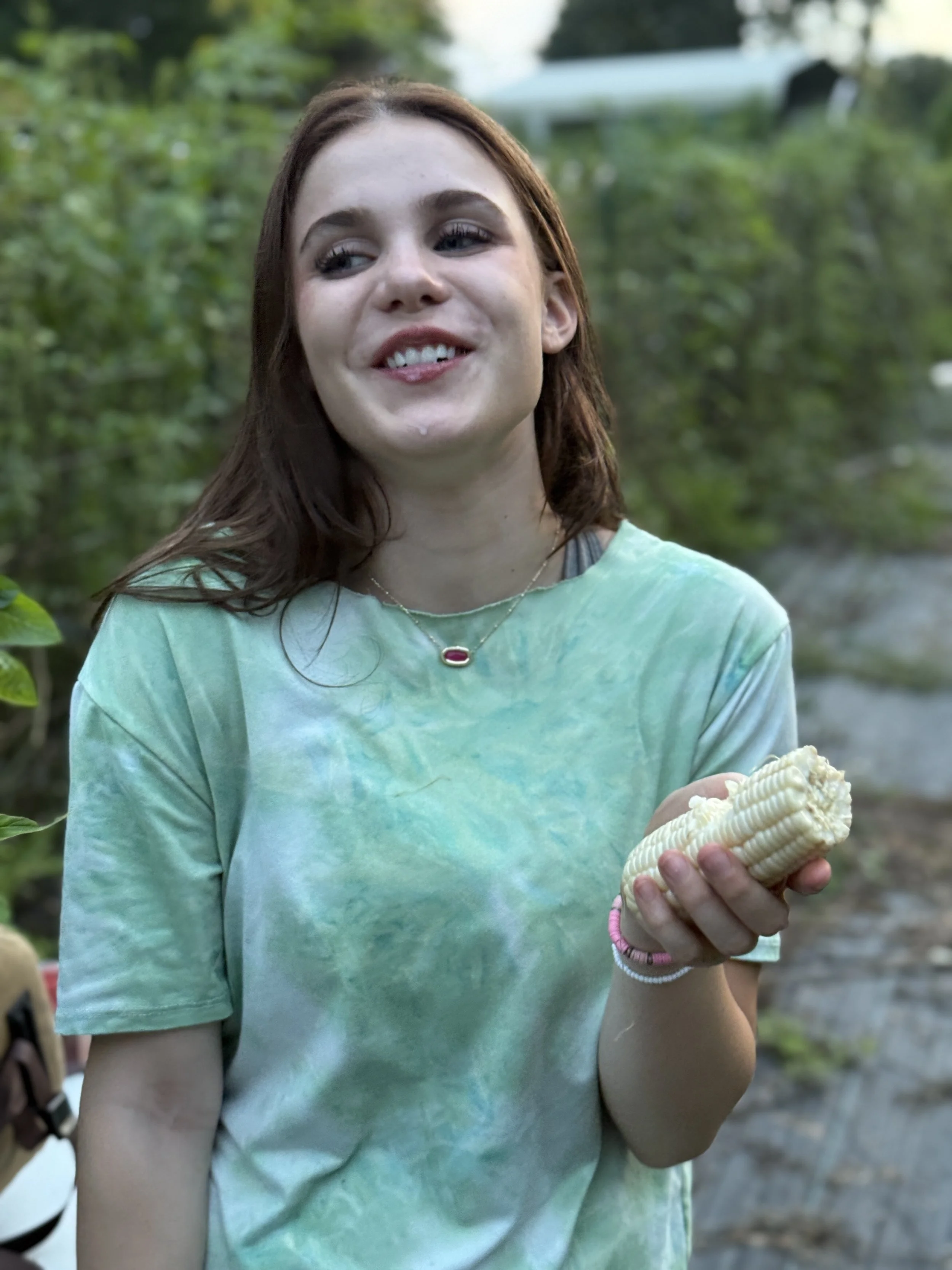 A young woman with shoulder-length brown hair smiling outdoors, holding an ear of corn, wearing a green tie-dye t-shirt and a beaded bracelet.