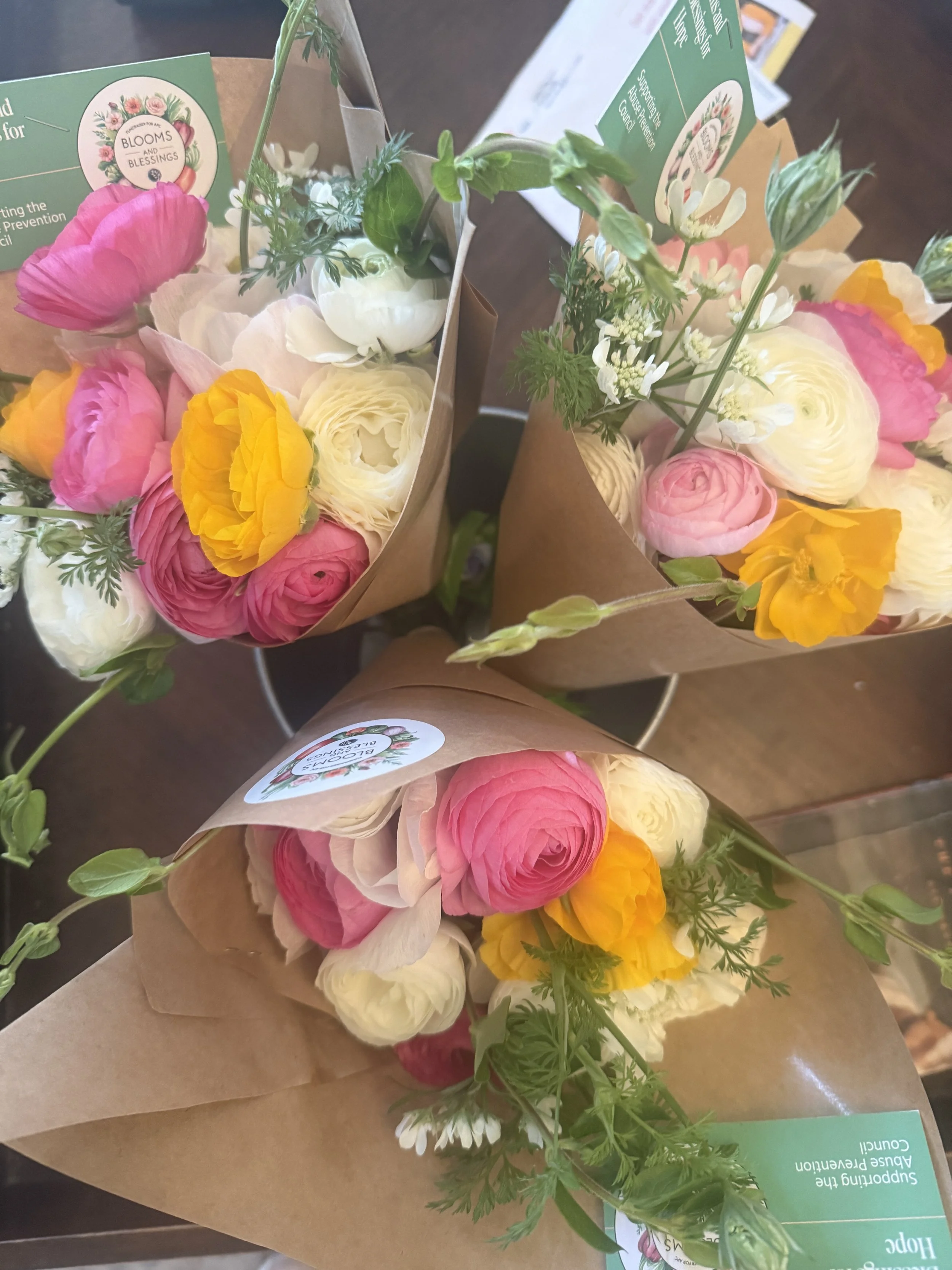 Three bouquets of flowers wrapped in brown paper with pink, white, and yellow blooms, along with green foliage, placed on a wooden surface.