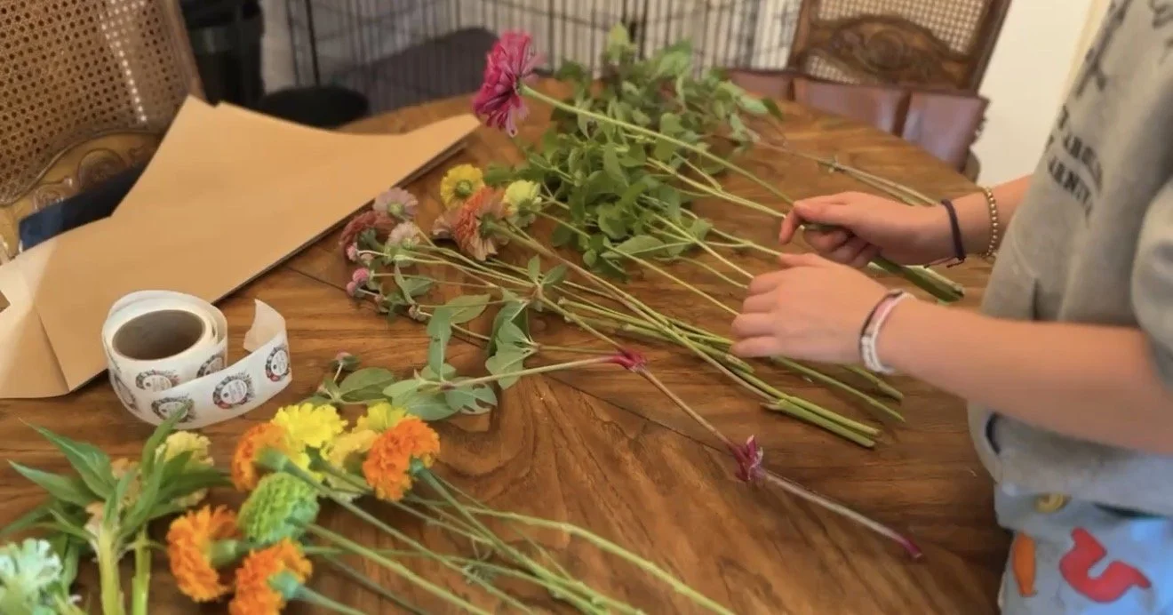 Person arranging colorful flowers on a wooden table with wrapping paper and tape nearby.