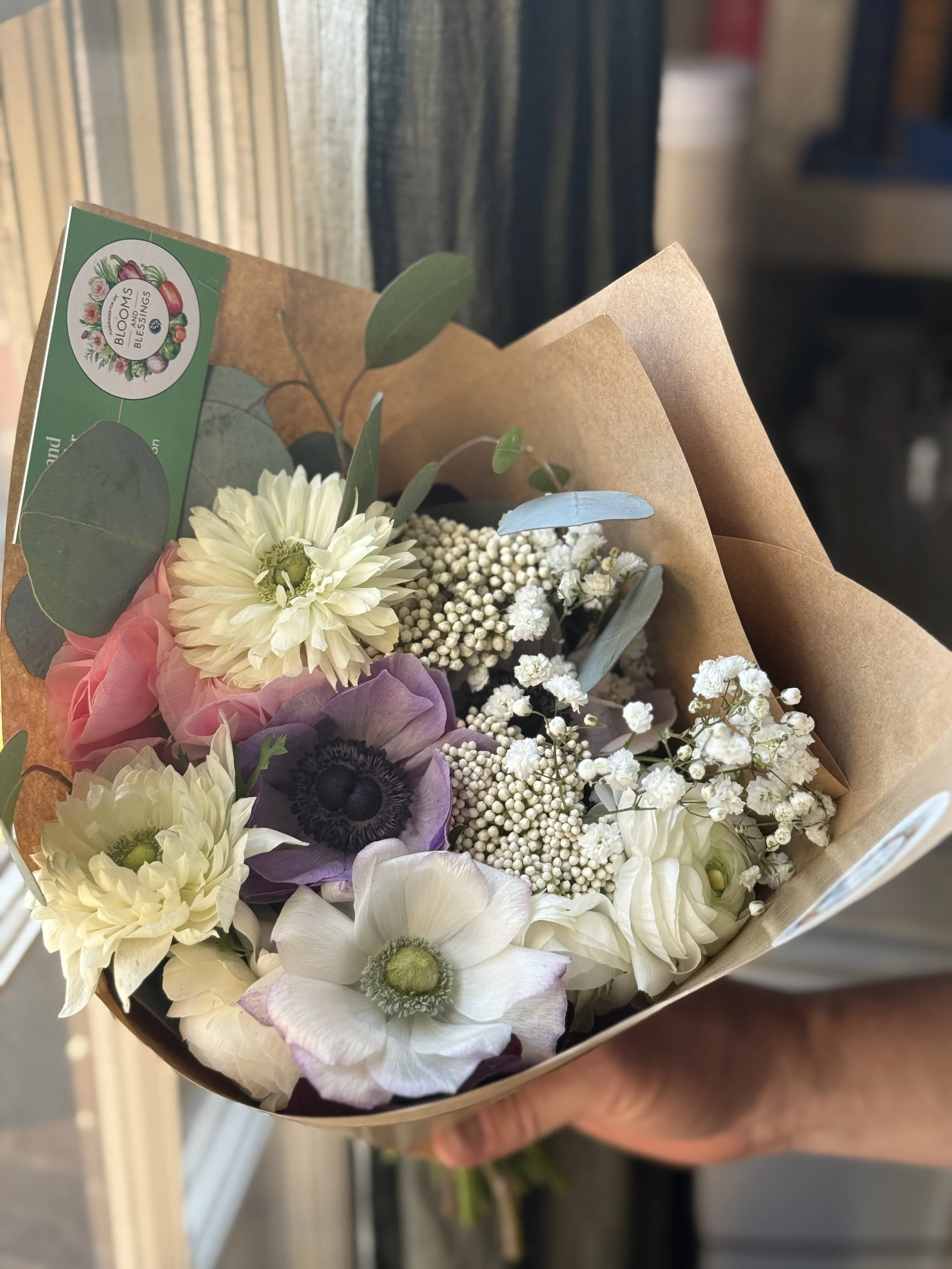 Hand holding a bouquet of mixed flowers including white, purple, and pink blooms with green leaves, wrapped in brown paper.