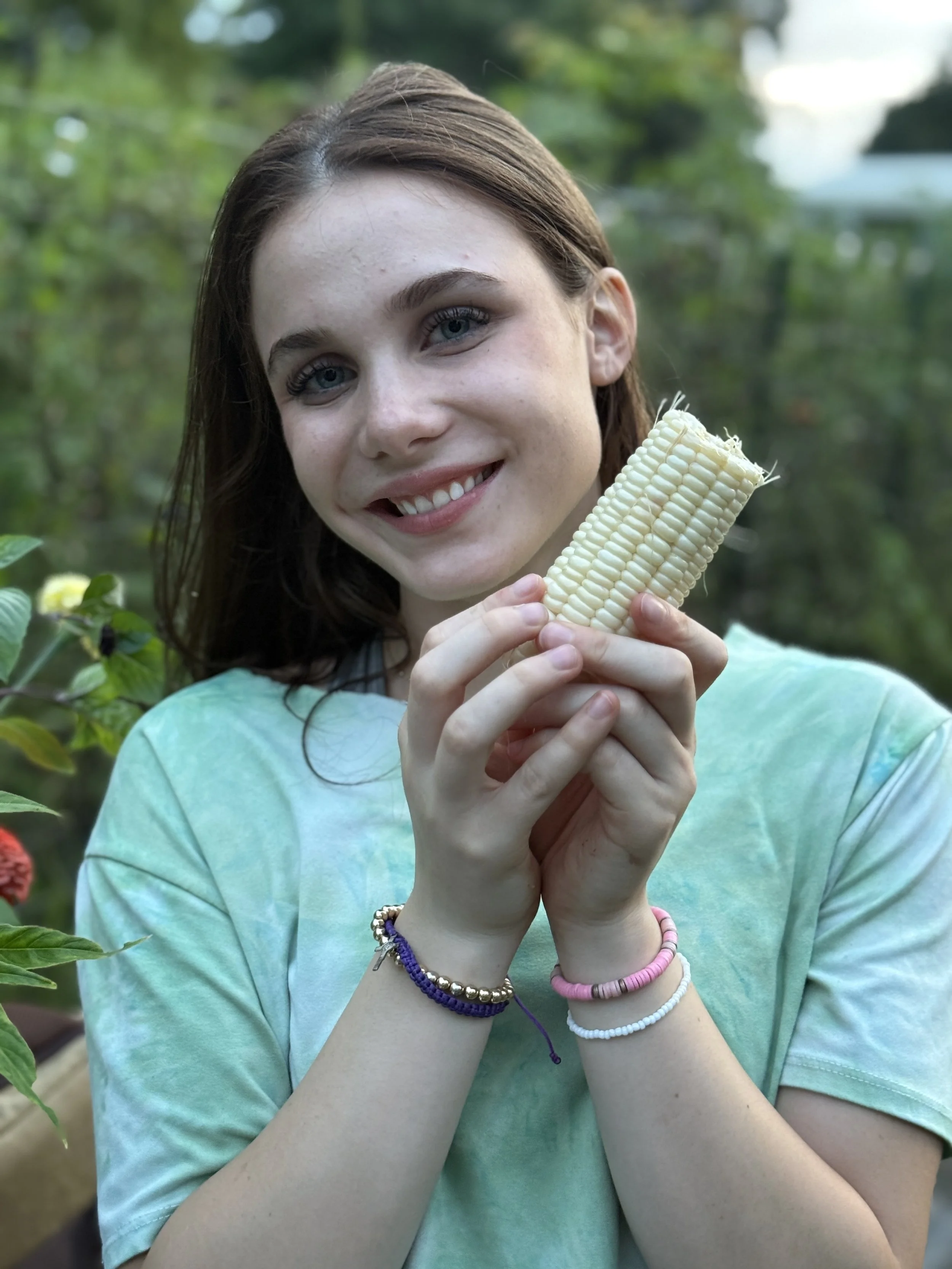 A smiling young woman with long brown hair holding a piece of corn on the cob outdoors in a garden or forest setting.