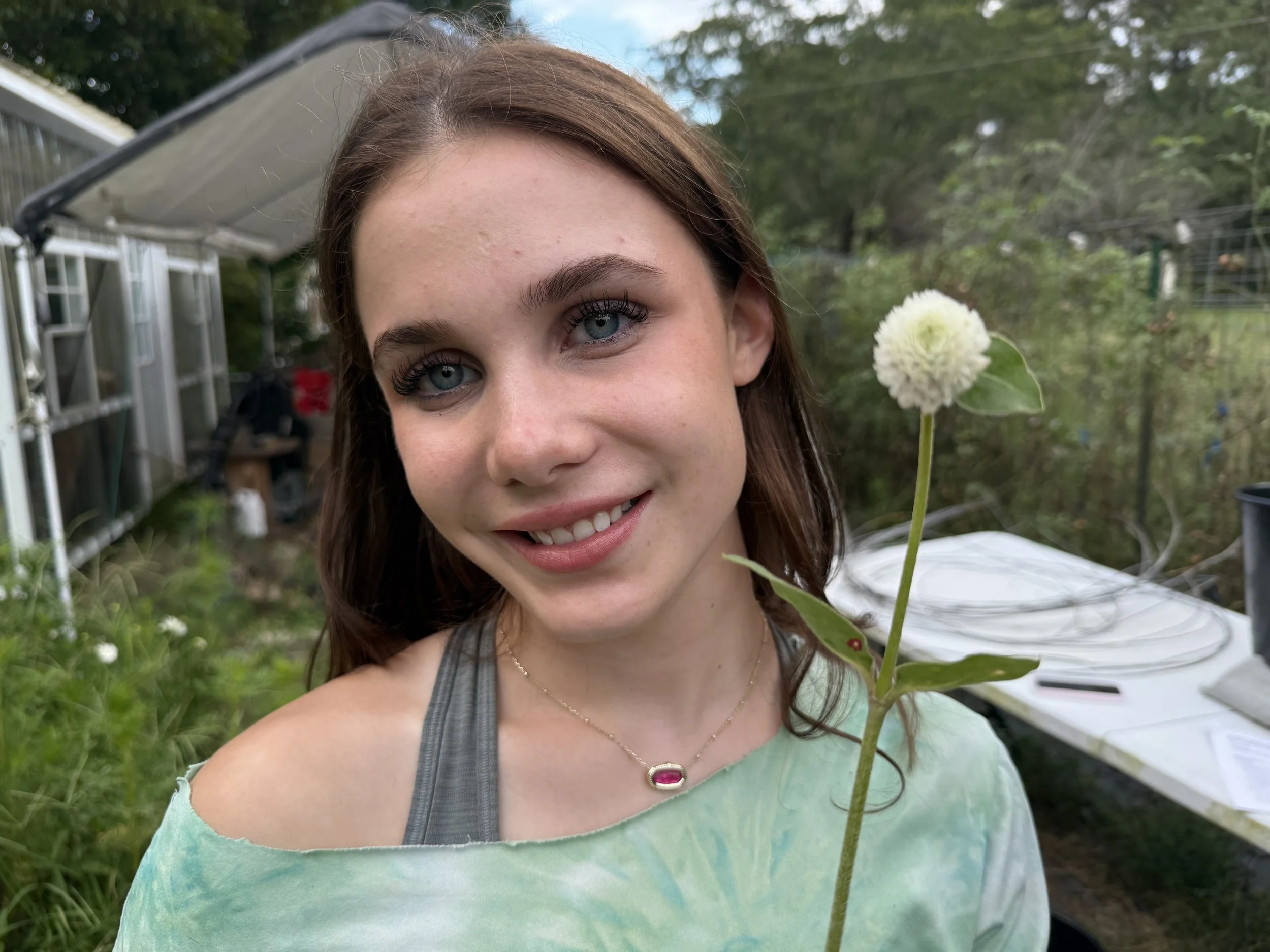 A young woman with blue eyes and brown hair smiling outdoors, holding a white pom-pom flower on a long stem with green leaves, in a garden setting with a greenhouse and trees in the background.