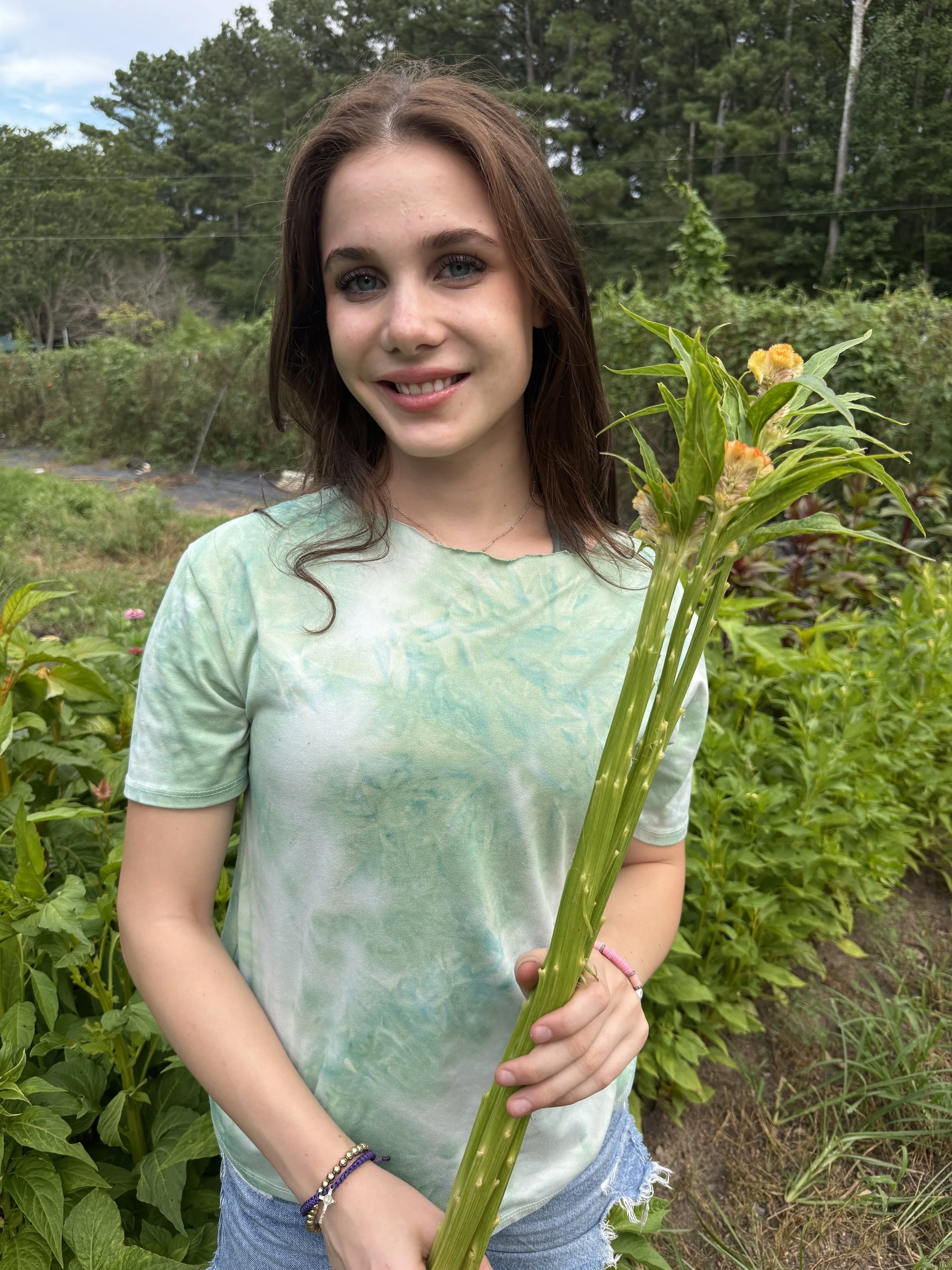 A young woman with brown hair and blue eyes smiling outdoors in a garden, holding a large green plant with yellow flowers.