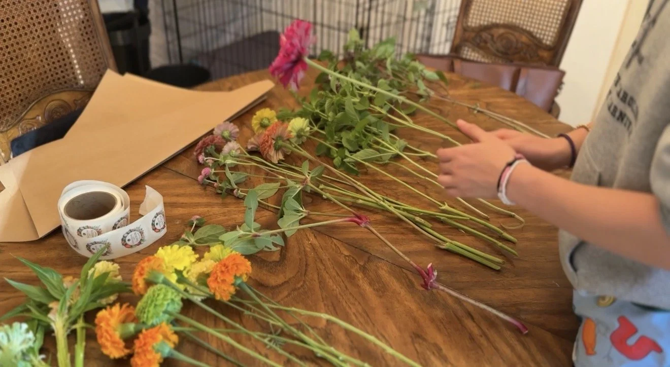 Person arranging pink, yellow, and orange flowers on a wooden table with wrapping paper and a cup of coffee nearby.