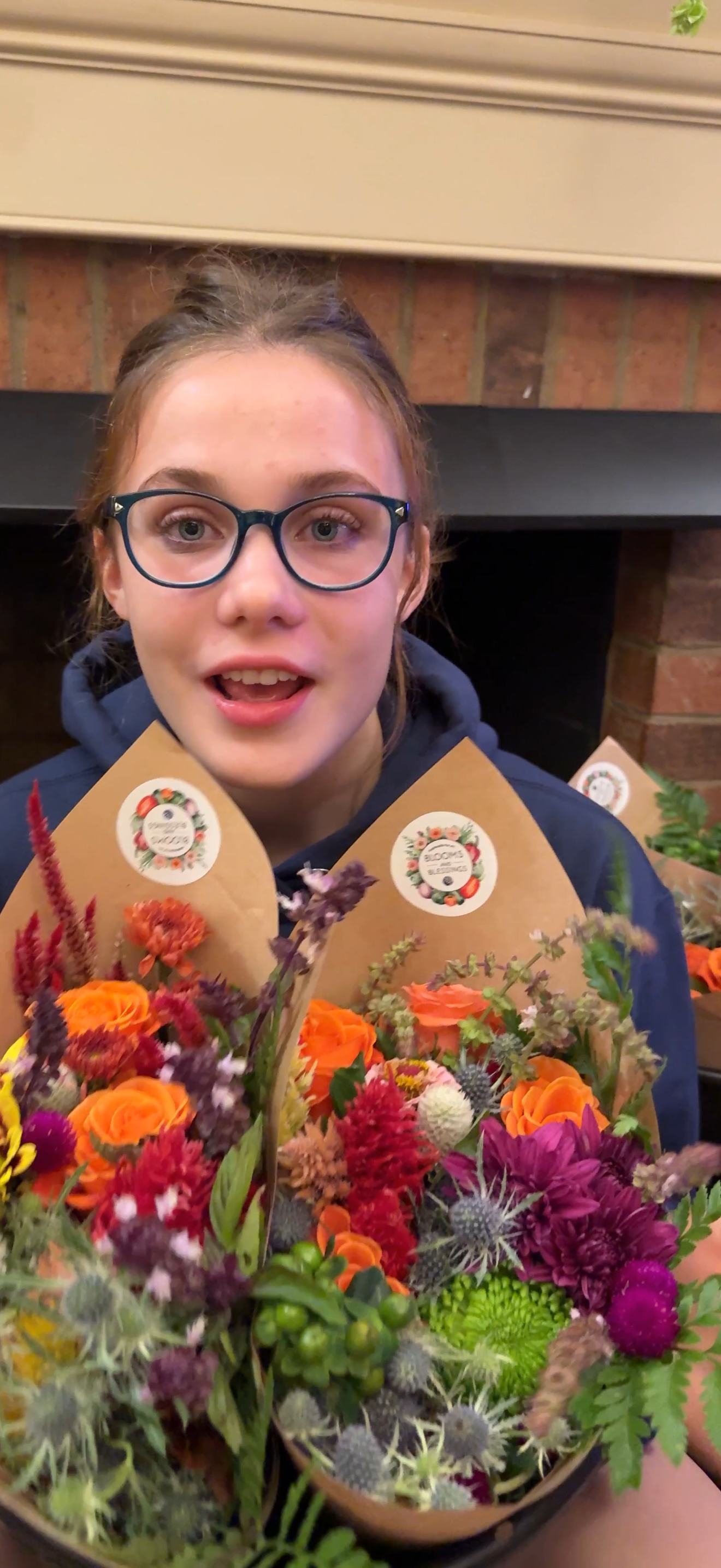 Young woman with glasses holding colorful flower bouquets with tags, standing in front of a brick fireplace.